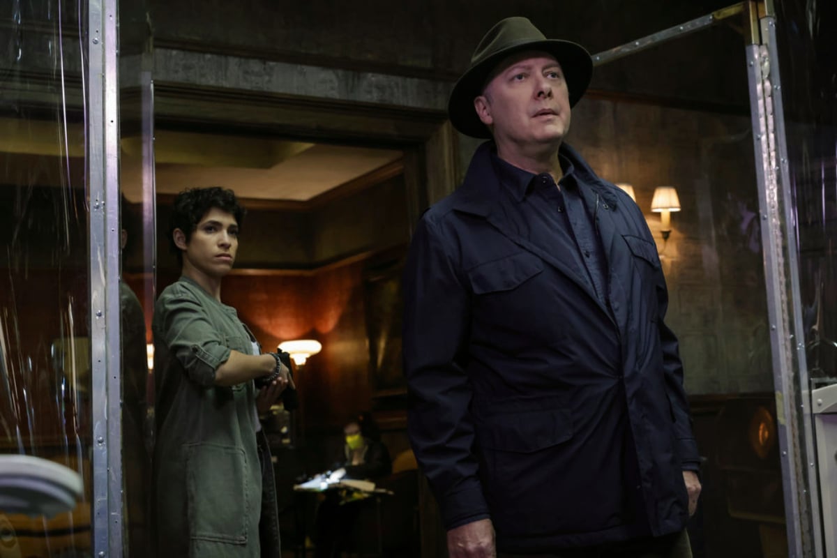 Diany Rodriguez as Weecha Xiu and James Spader as Red in The Blacklist Season 9. Weecha stands behind Red, who is wearing a dark coat and hat.