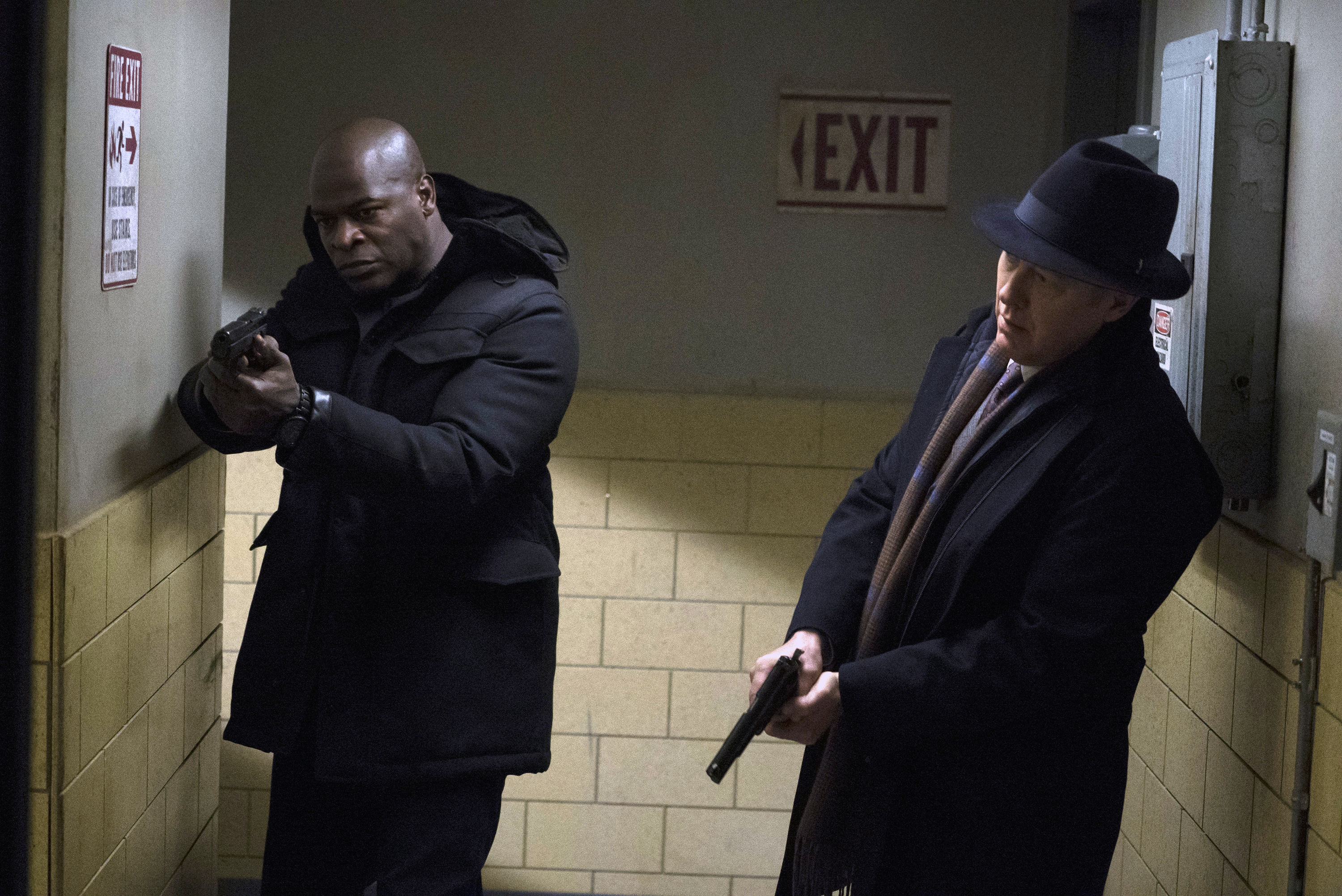 'The Blacklist' Season 9 stars Hisham Tawfiq and James Spader, in character as Dembe and Red, wield their guns. Dembe wears a black winter coat and black pants. Red wears a black dress coat and a black fedora.