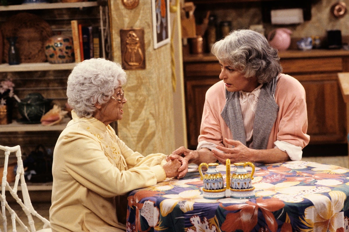'The Golden Girls' actor Estelle Getty in a yellow robe, and Bea Arthur in a pink, white, and grey nightgown; sitting at a kitchen table.