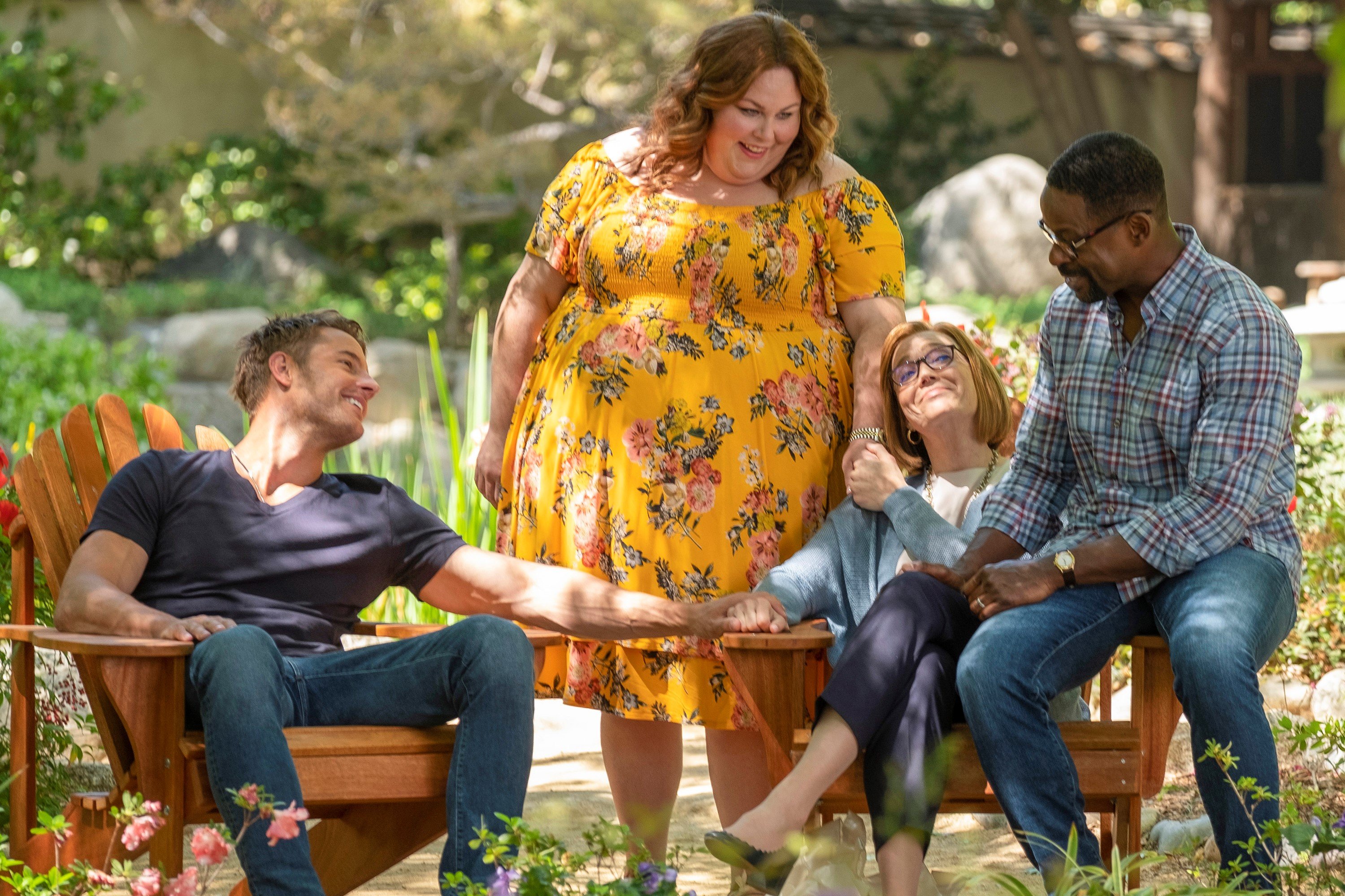 'This Is Us' Season 6 stars Justin Hartley, Chrissy Metz, Mandy Moore, and Sterling K. Brown, in character as Kevin, Kate, Rebecca, and Randall, hold hands and sit outside. Kevin wears a dark blue shirt with jeans. Kate wears a yellow dress with flowers on it. Rebecca wears a light blue cardigan and dark blue pants. Randall wears a plaid button-up shirt and jeans. 'This Is Us' returns with season 6 on Jan 4.