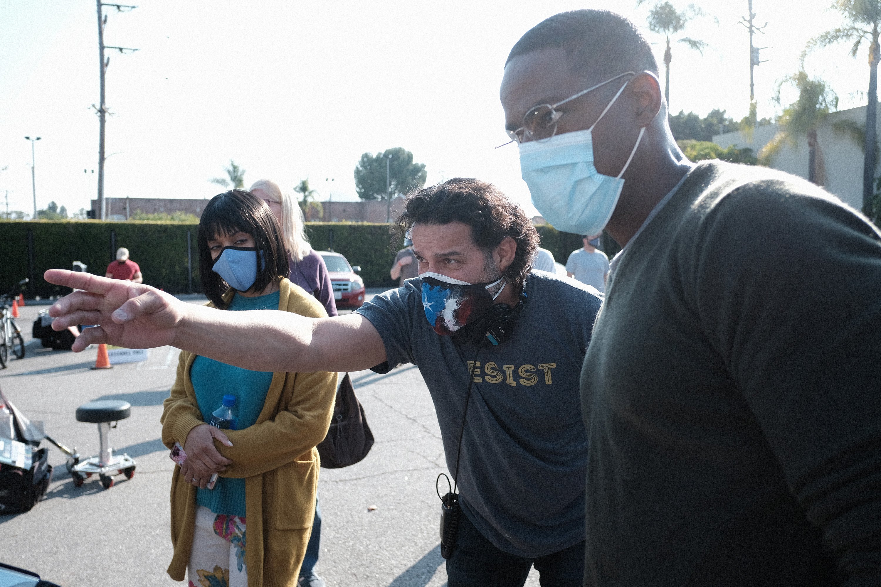 'This Is Us' Season 6 stars Susan Kelechi Watson, Jon Huertas, and Sterling K. Brown on set in season 5. Huertas directs a scene between Kelechi Watson and Brown. Kelechi Watson wears a bob wig with bangs, a yellow cardigan over a teal shirt and floral pants. Huertas wears a gray shirt that says, 'Resist.' Brown wears a dark sweater and glasses. All three stars are wearing masks.