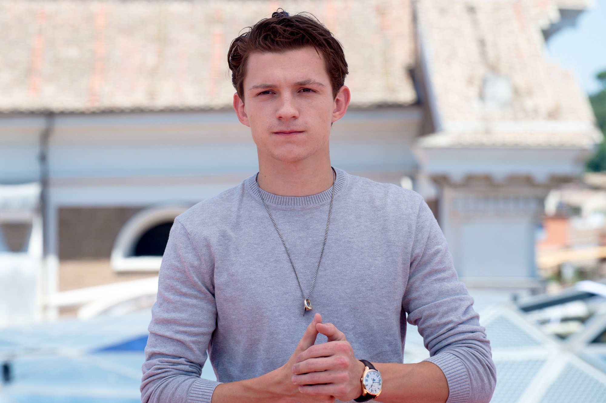 Spider-Man actor Tom Holland wearing a grey, long-sleeved shirt and a watch. He's standing with his hands clasped together.