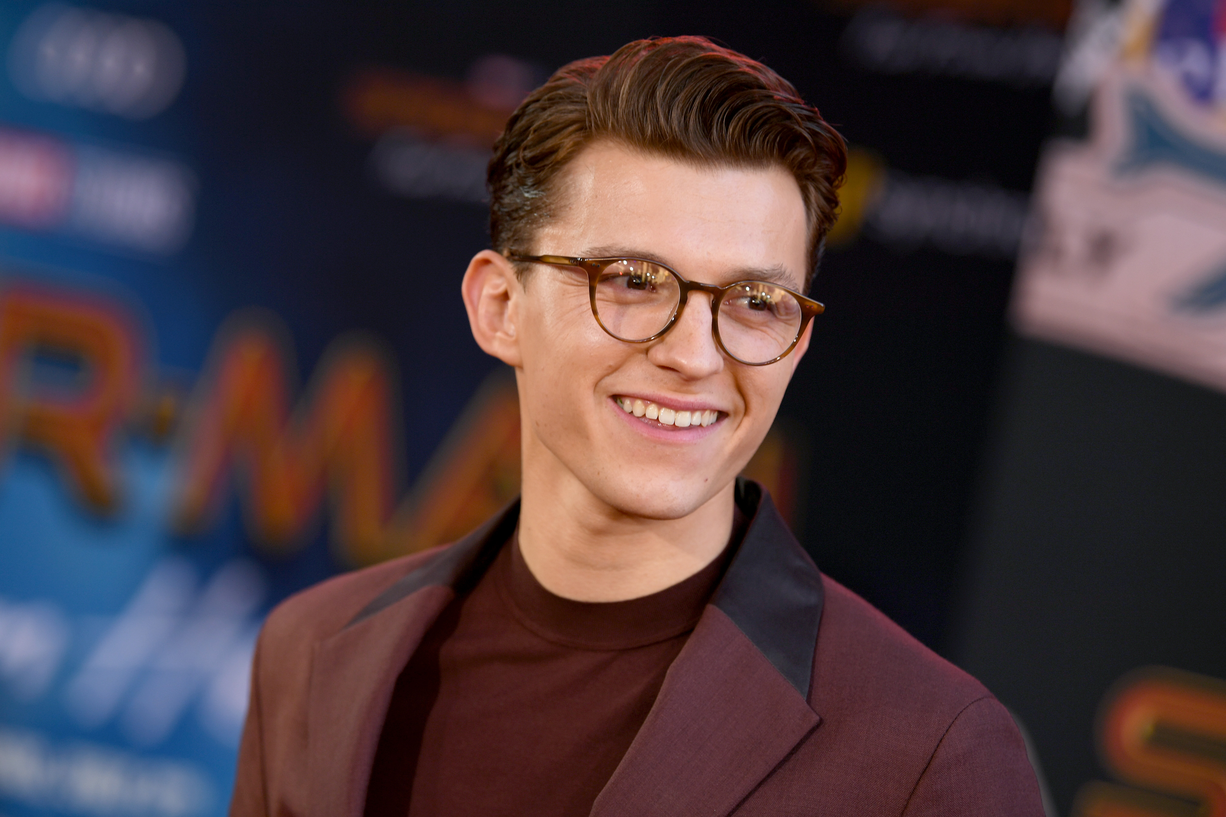 'Spider-Man: No Way Home' star Tom Holland wears a maroon suit over a darker maroon t-shirt and wears a pair of glasses.