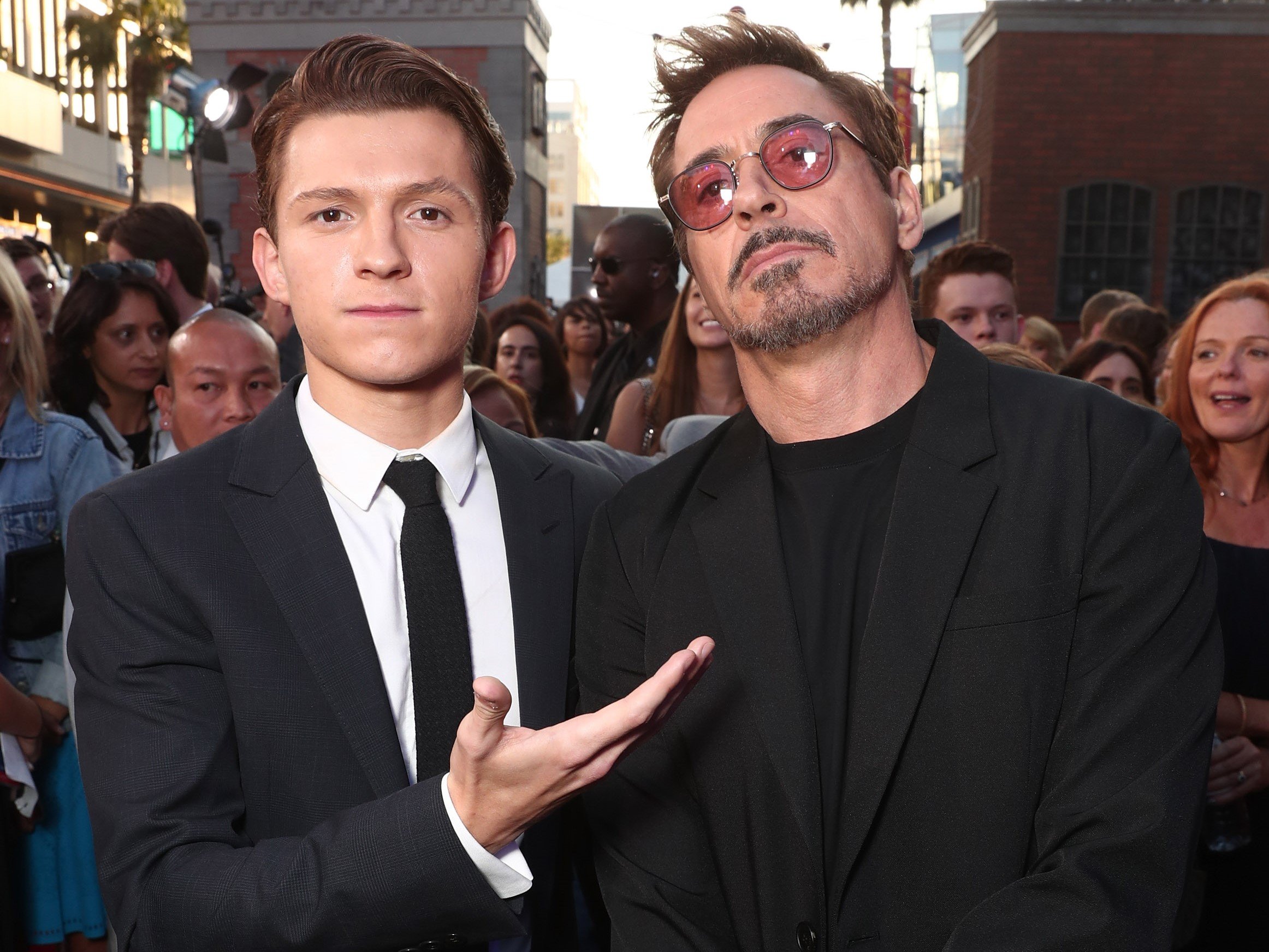 'Spider-Man' stars Tom Holland and Robert Downey Jr. pose for a picture. Holland wears a black suit over a white shirt and black tie. Downey Jr. wears a black suit over a black shirt and red-tinted glasses.