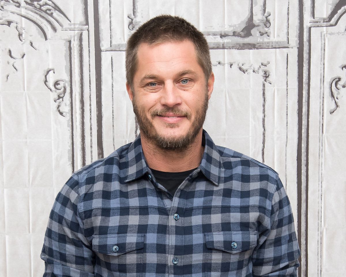 Travis Fimmel smiles in a plaid shirt, in front of a white background
