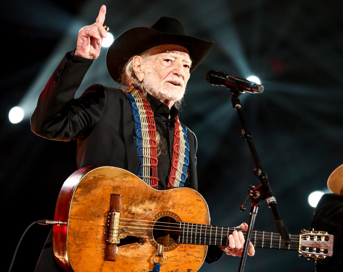 Willie Nelson performs with his acoustic guitar, dressed in black and wearing a cowboy hat, holding one finger in the air