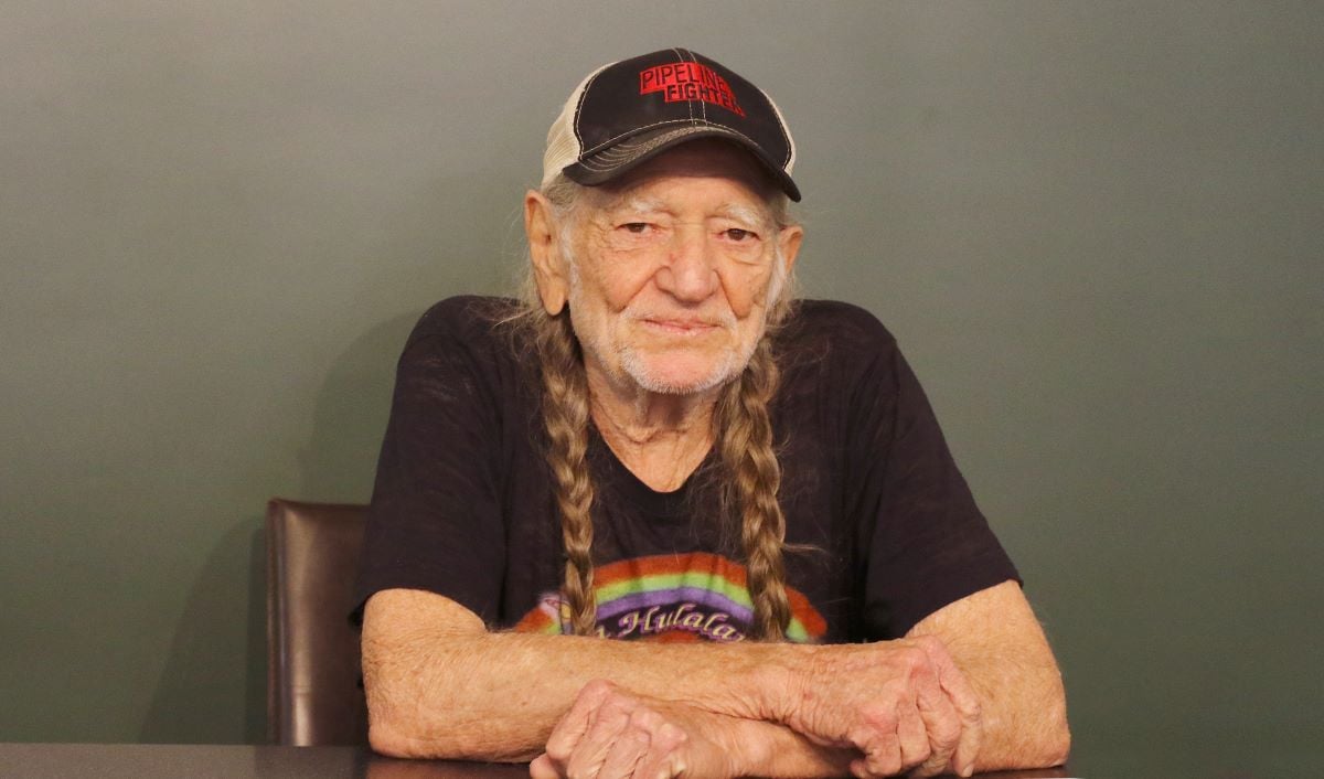 Willie Nelson wearing a cap over his braids, seated behind a table in a tee shirt