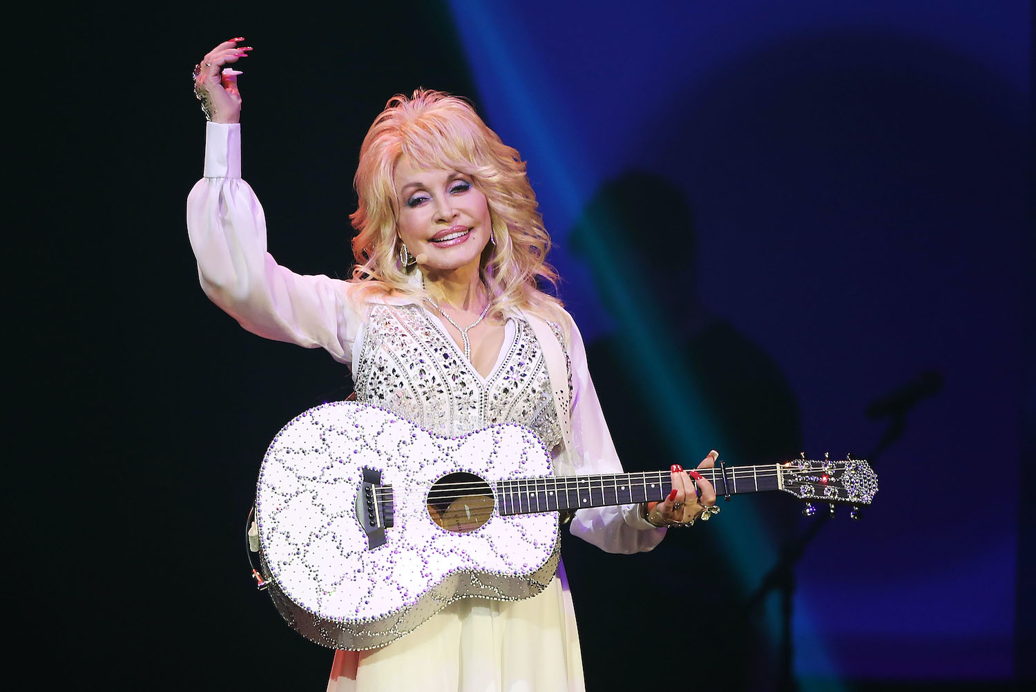Dolly Parton wears a white dress while holding a white jeweled guitar and smiling with one hand in the air during a performance in Sydney, Australia