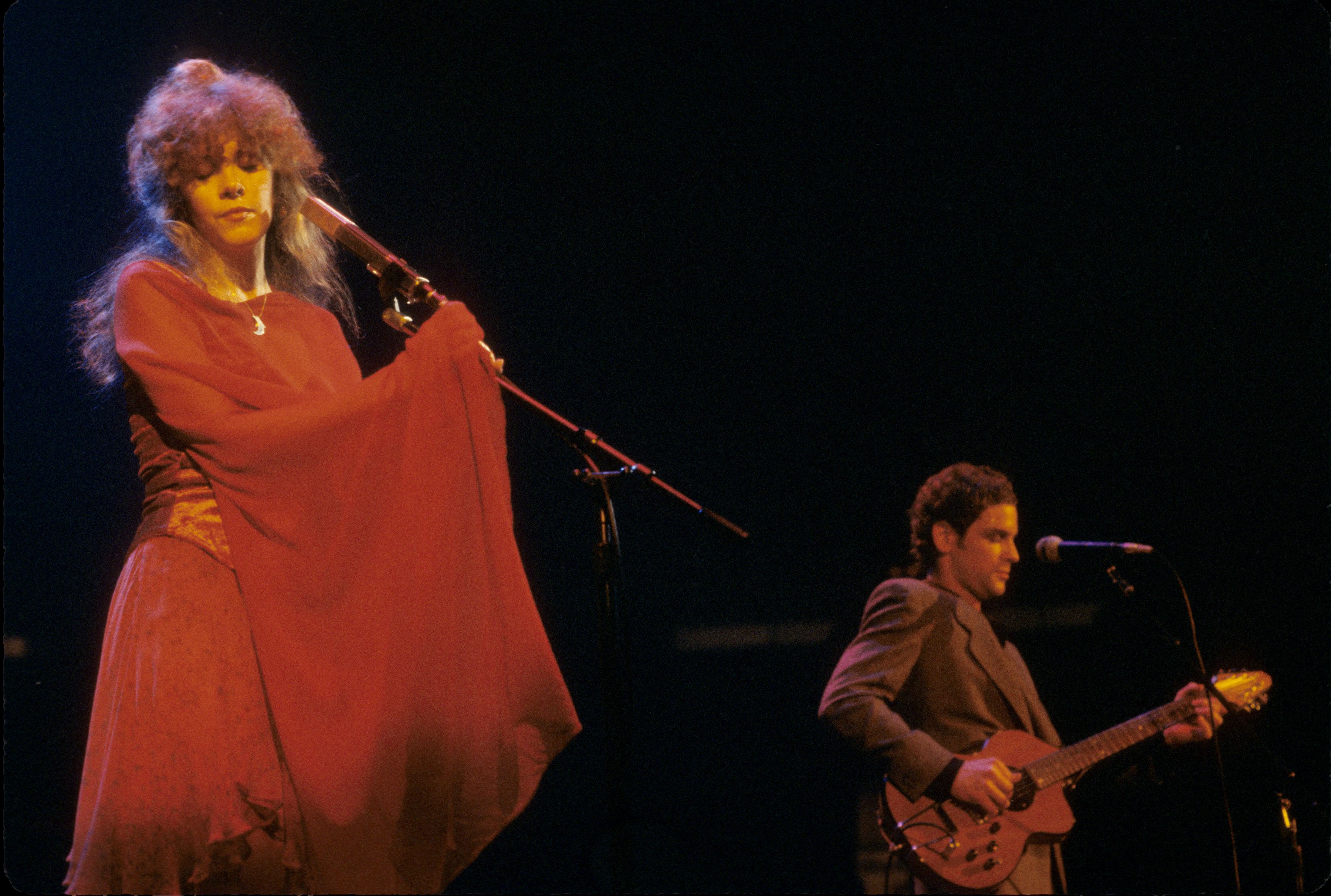 Stevie Nicks wears a long red dress and holds a microphone and Lindsey Buckingham plays guitar.
