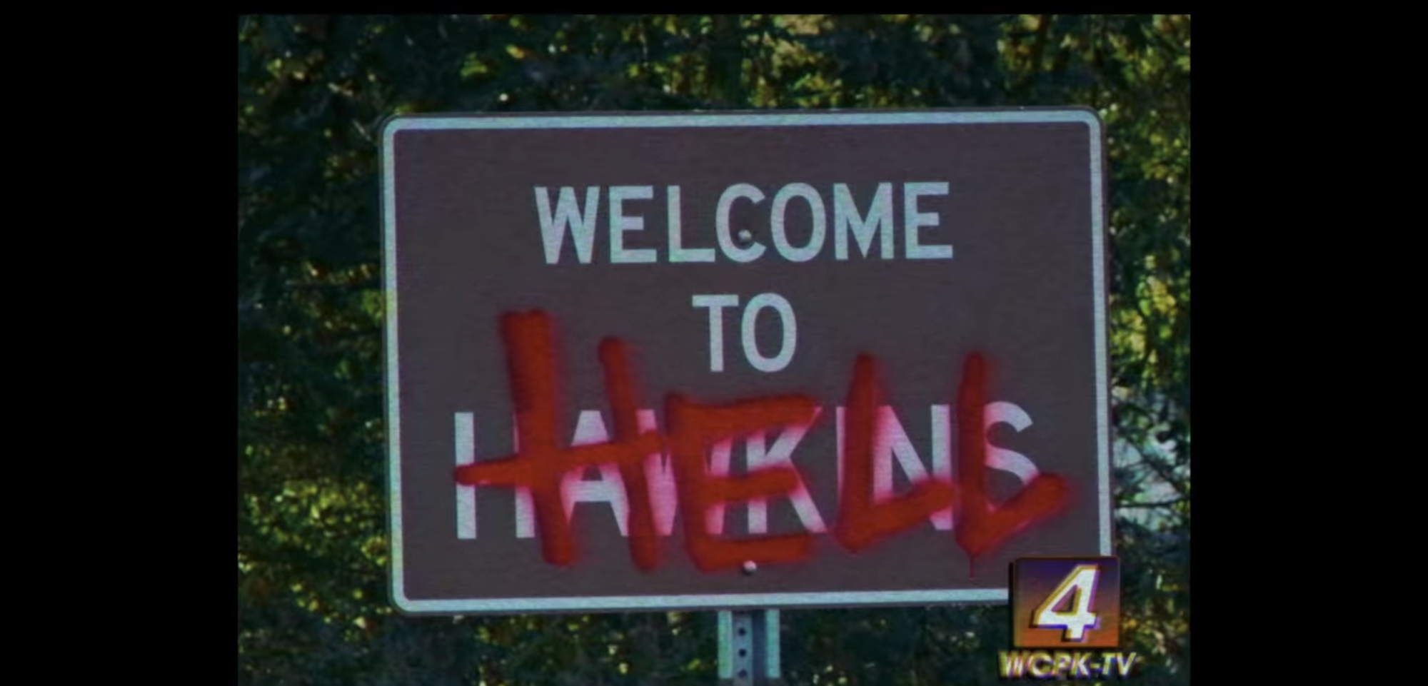 A road sign saying "Welcome to Hawkins" with the word "Hell" spray painted over the word "Hawkins" in a scene from 'Stranger Things' Season 3