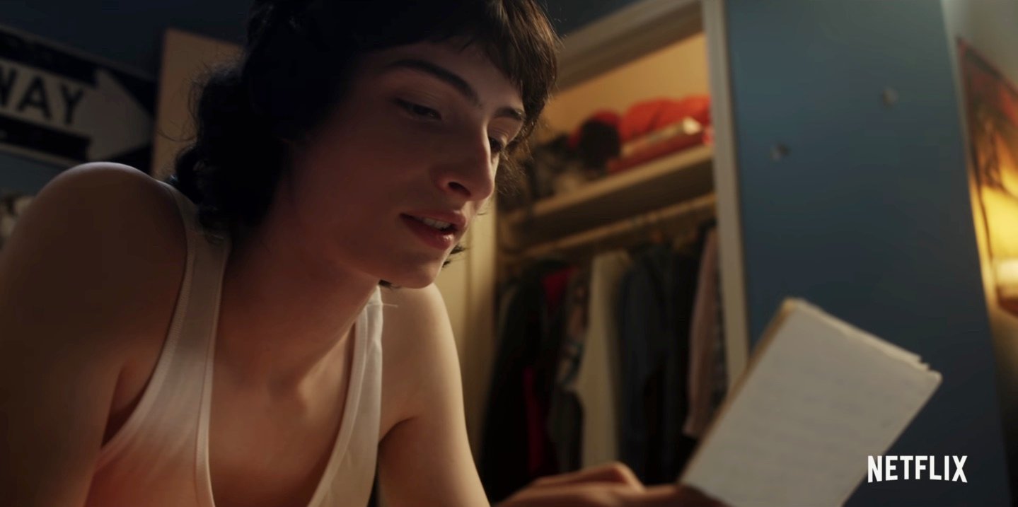 A still from 'Stranger Things' Season 4 with Mike Wheeler, played by Finn Wolfhard, in a white tank top reading a letter.