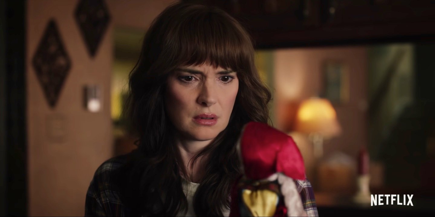 Winona Ryder as Joyce Byers wearing a flannel shirt and staring at a Russian doll in a still from 'Stranger Things' Season 4