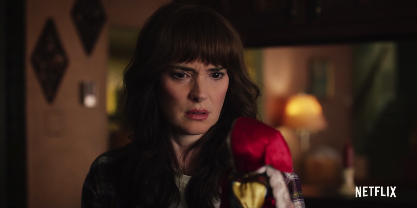 Winona Ryder as Joyce Byers wearing a flannel shirt and staring at a Russian doll in a still from 'Stranger Things' Season 4