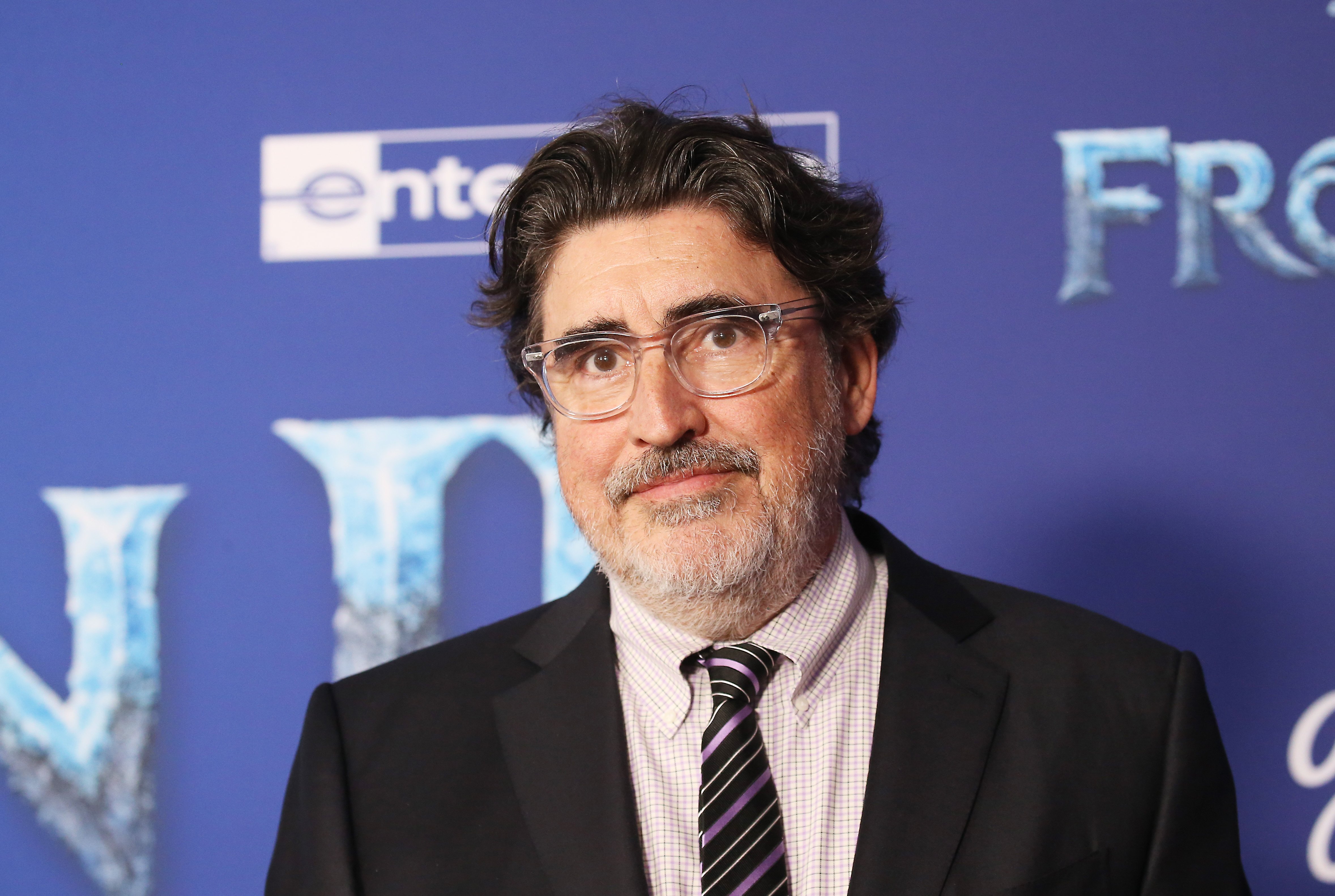 'Spider-Man: No Way Home' star Alfred Molina wears a black suit over a white checkered button-up shirt and a black tie with purple and white stripes.