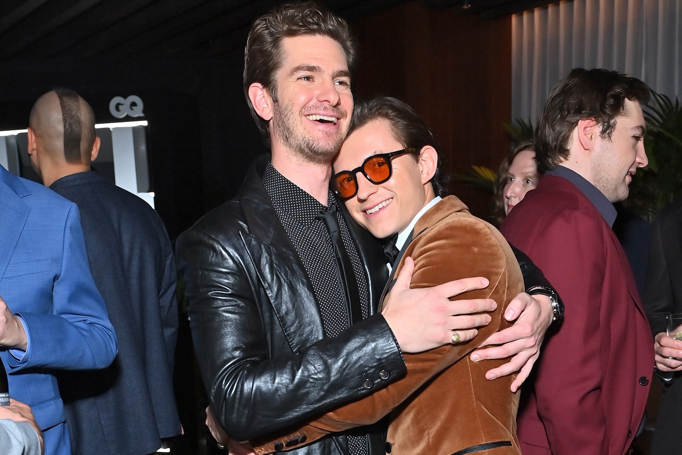 'Spider-Man: No Way Home' star Tom Holland embraces Andrew Garfield. Holland wears a light brown velvet suit jacket and orange-tinted glasses. Garfield wears a black leather jacket over a black button-up shirt with white polka dots and a black tie. Andrew Garfield, along with Tobey Maguire, are rumored to appear in 'Spider-Man: No Way Home.'