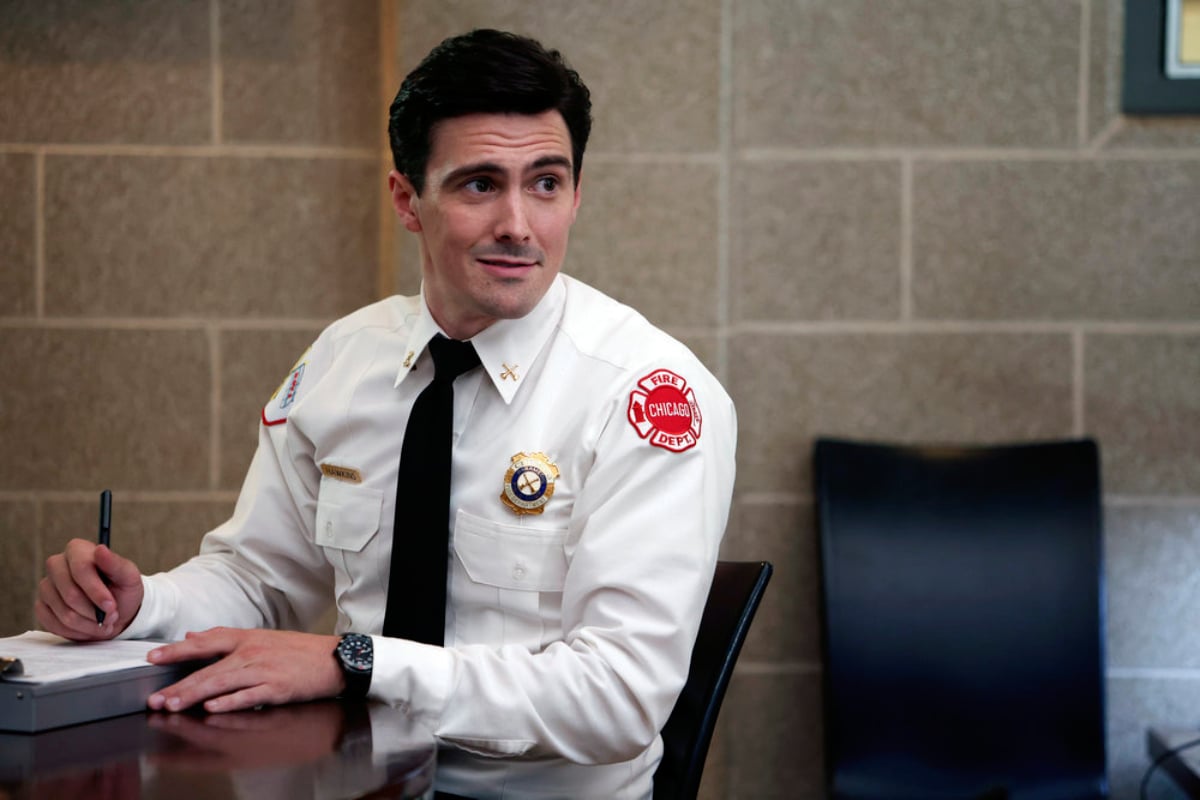 Jimmy Nicholas as Chief Hawkins in Chicago Fire Season 10. Hawkins sits at a desk with a pen and paper. He is wearing a white shirt and a tie and smiling.