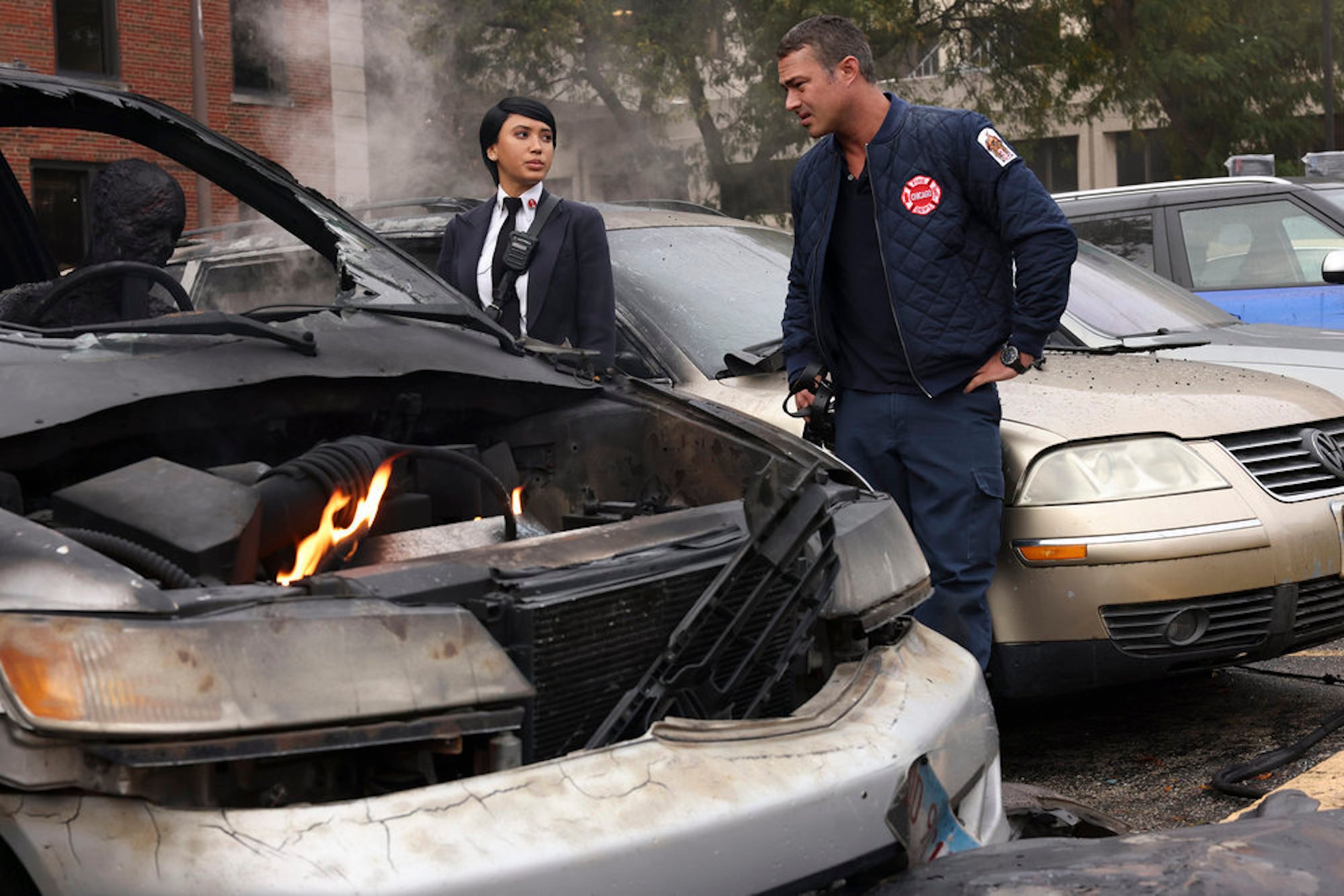 Andy Allo as Wendy Seager and Taylor Kinney as Kelly Severide standing next to a car on fire in 'Chicago Fire' Season 10