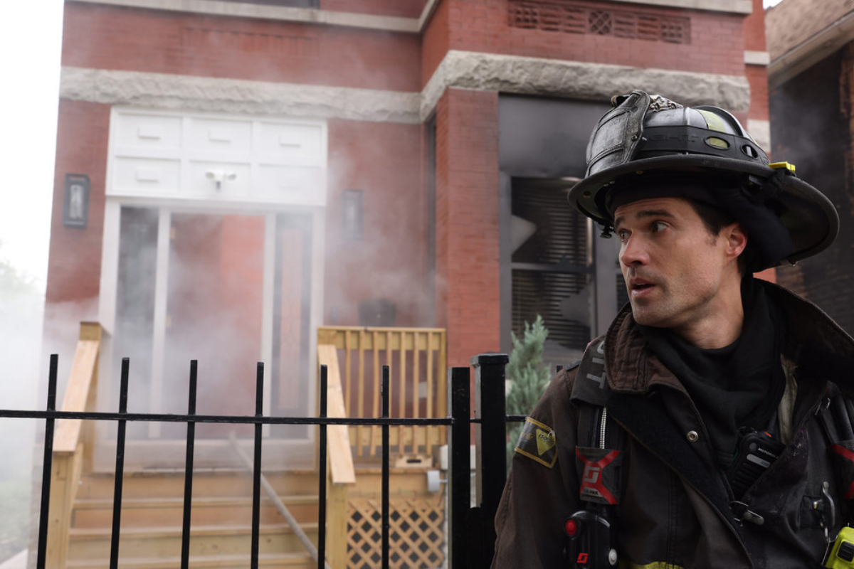 Brett Dalton at Jason Pelham in Chicago Fire Season 10. Pelham stands in front of a smoking building in firefighter gear.