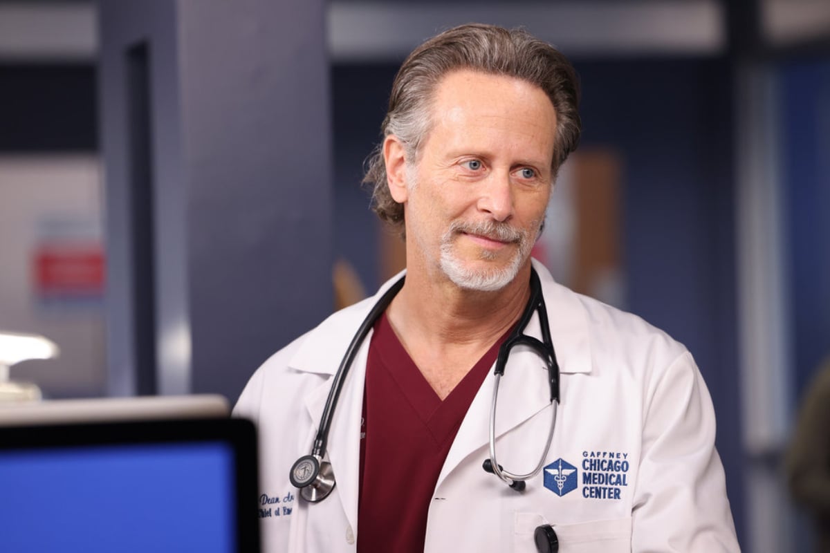 Steven Weber as Dr. Dean Archer in Chicago Med Season 7. Archer is wearing a white lab coat, red scrubs, and a stethoscope around his neck.