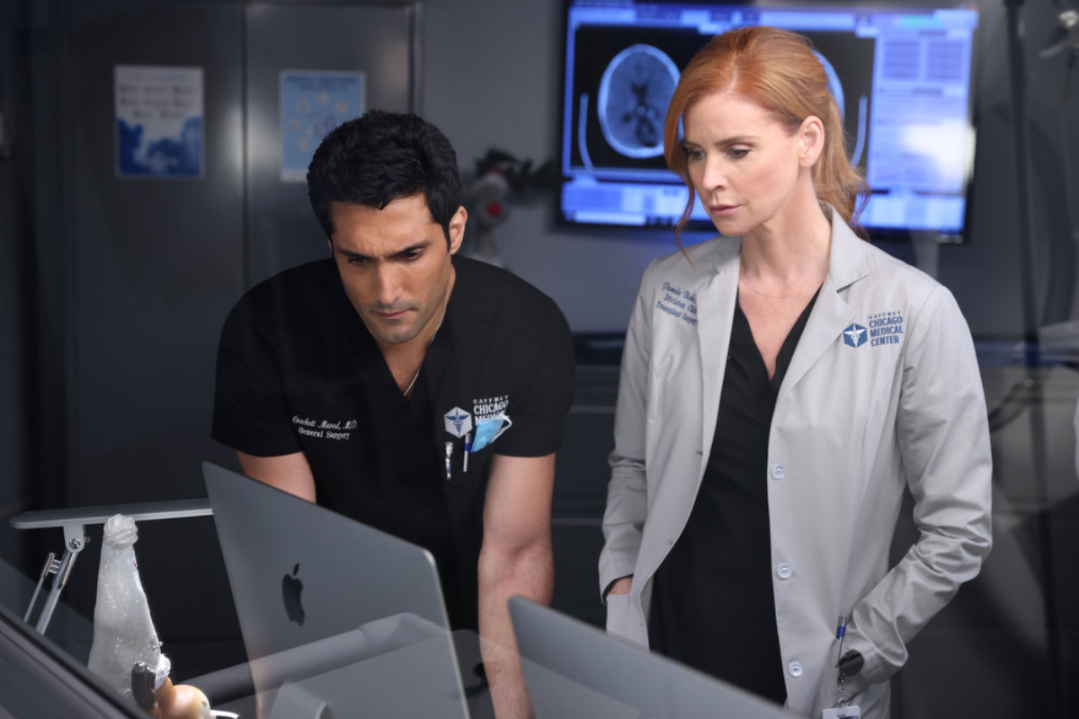 Dominic Rains as Crockett Marcel and Sarah Rafferty as Dr. Pamela Blake in Chicago Med Season 7. Blake and Crockett lean over a computer with pictures of brain scans behind them. 