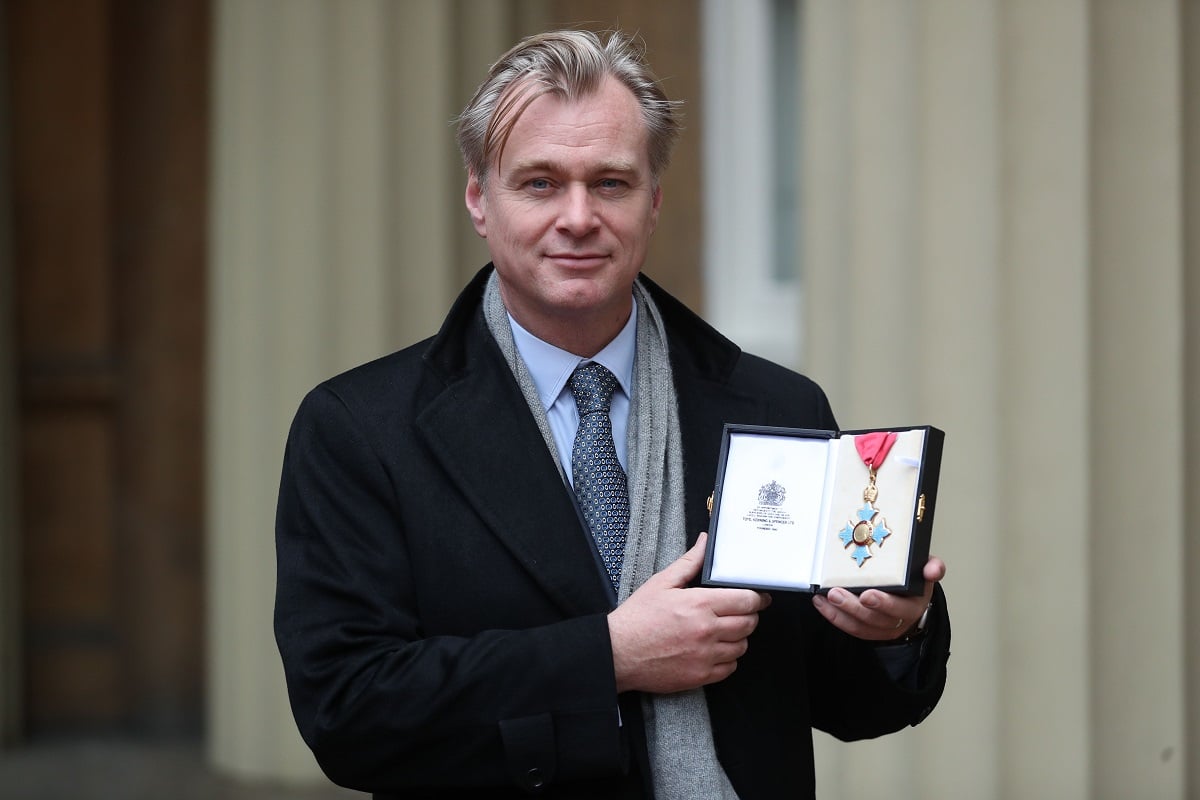 Christopher Nolan smirking while holding an award and wearing a trench coat.