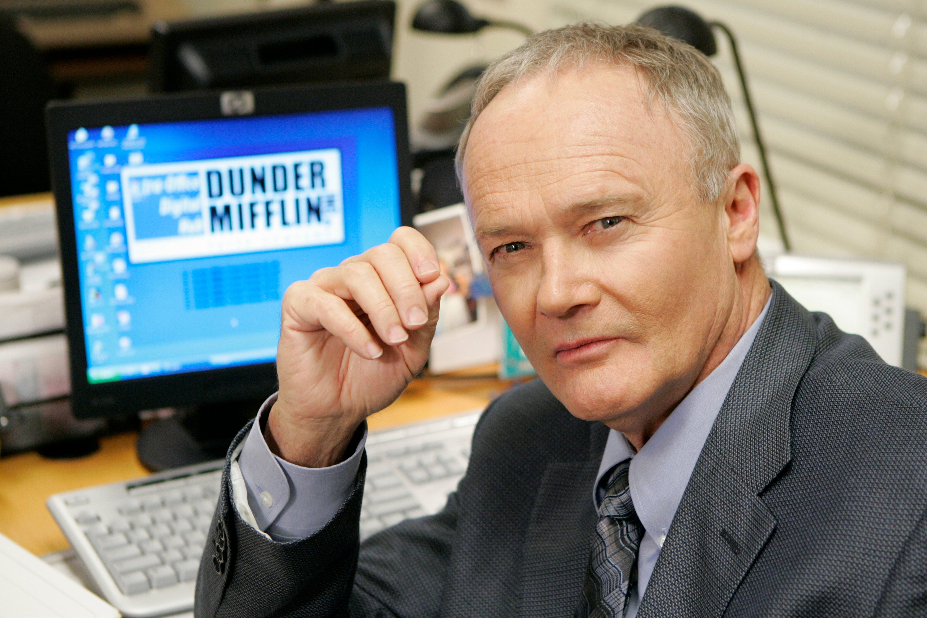 Creed Bratton as Creed Bratton on 'The Office' at a desk in front of a computer that has a Dunder Mifflin screen