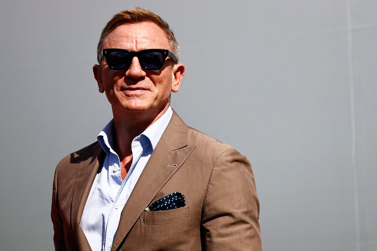 Daniel Craig posing in a brown suit wearing sunglasses.