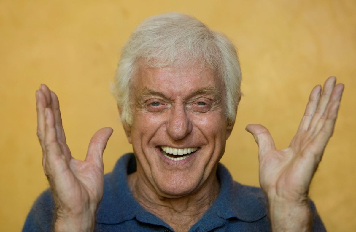 Dick Van Dyke smiles with his hands up, in a blue collared shirt in front of a yellow background