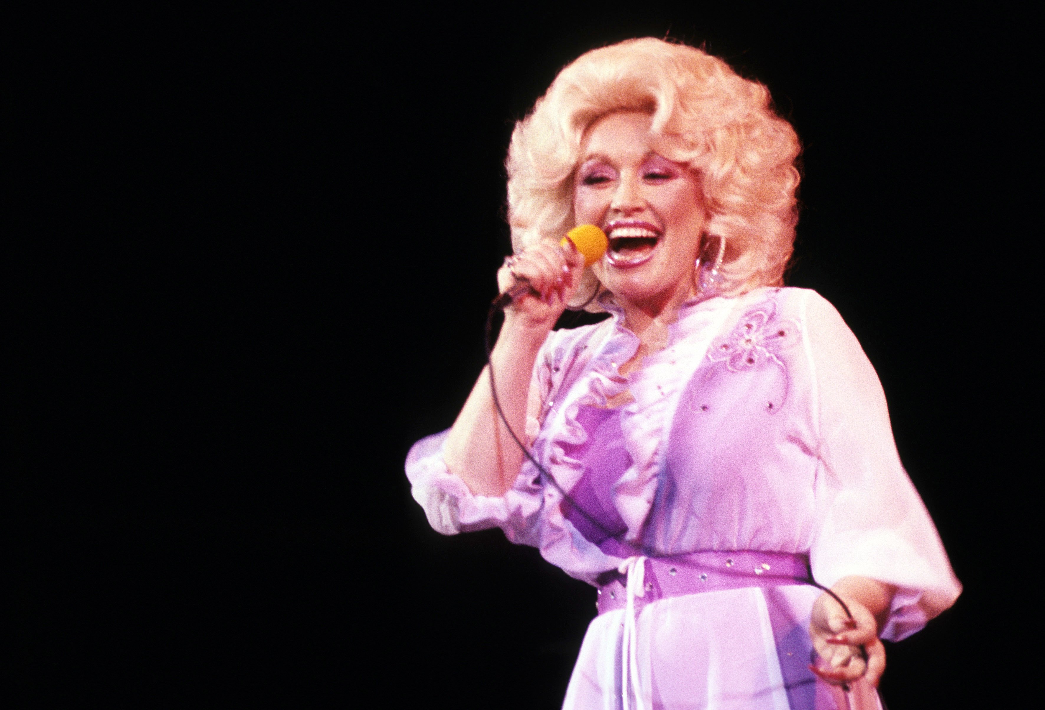 Dolly Parton wears a purple dress and sings into a microphone. 