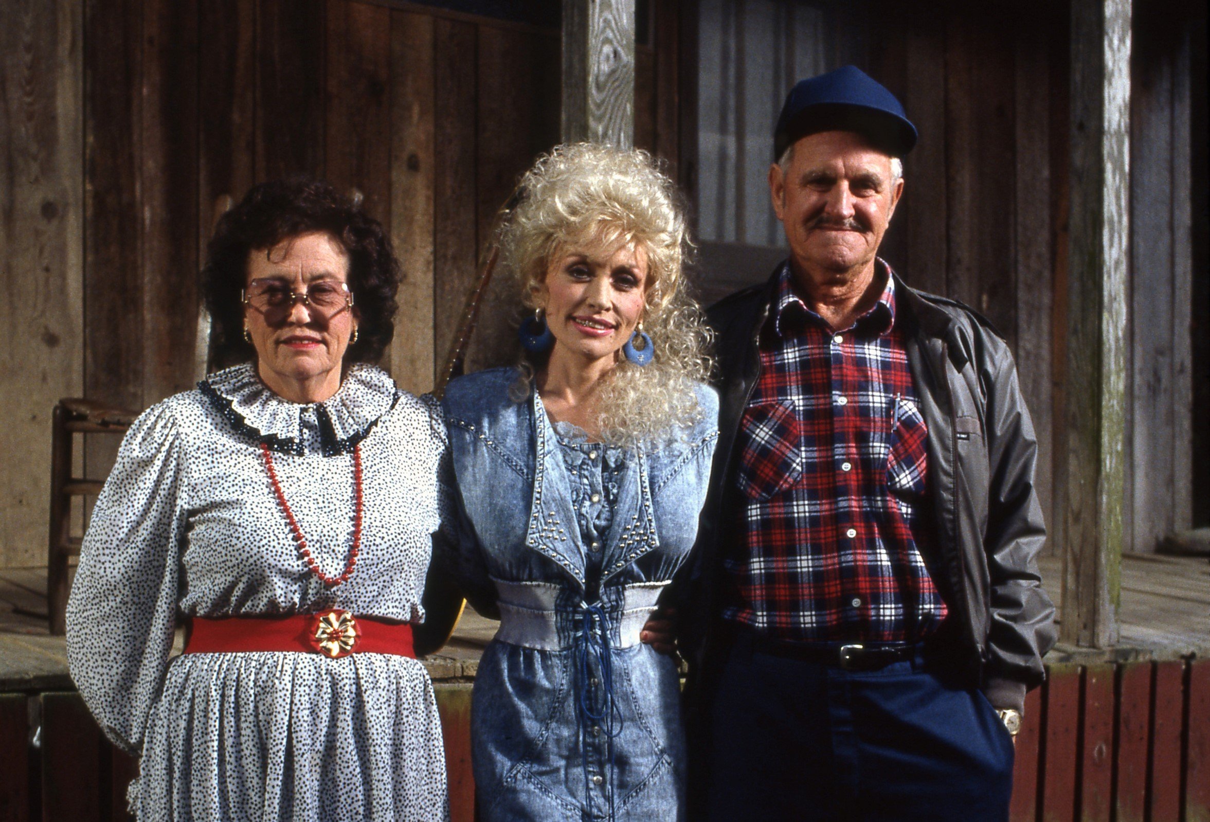 Dolly Parton's mother Avie Lee Owens wears a black and white dress with a red belt. Dolly Parton wears a blue dress. Dolly Parton's father Lee Parton wears a red plaid shirt and blue hat.