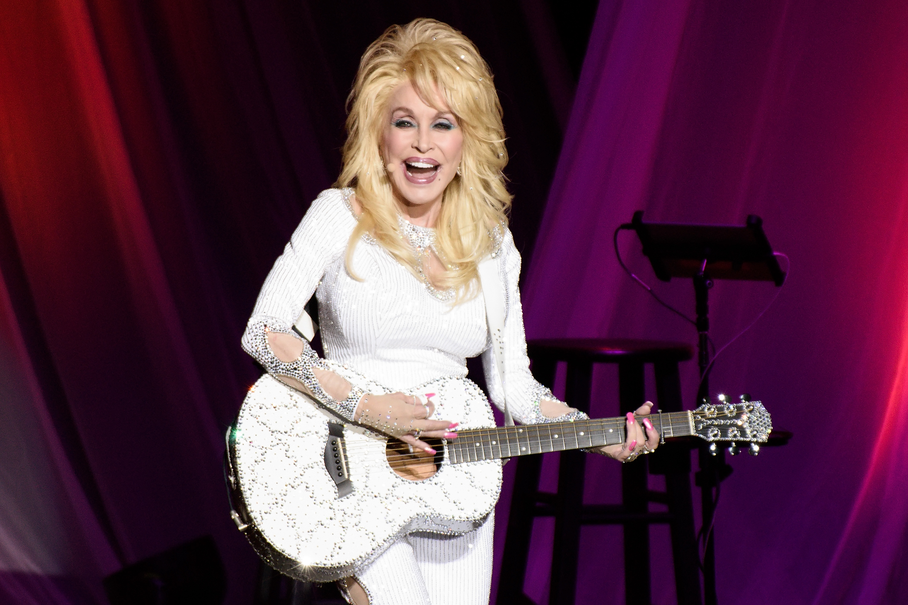 Dolly Parton, smiling and looking happy, wears an all white outfit and carries a white guitar.