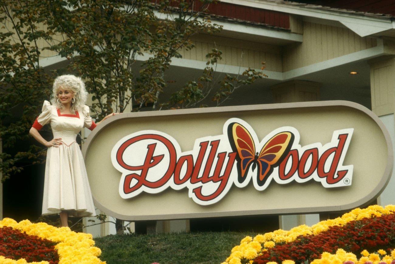 Dolly Parton wears a white dress and stands outside in front of a Dollywood sign.
