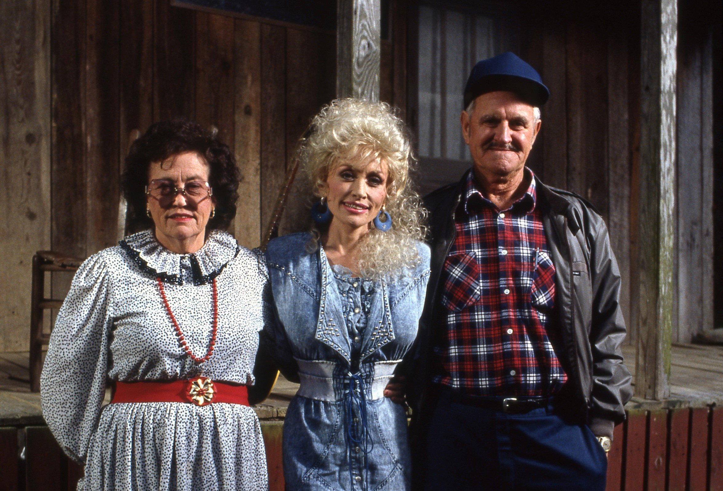 Dolly Parton's mother wears a black and white dress with a red belt. Dolly Parton wears a denim dress. Dolly Parton's father wears a red plaid shirt and blue hat. They stand in front of a house.