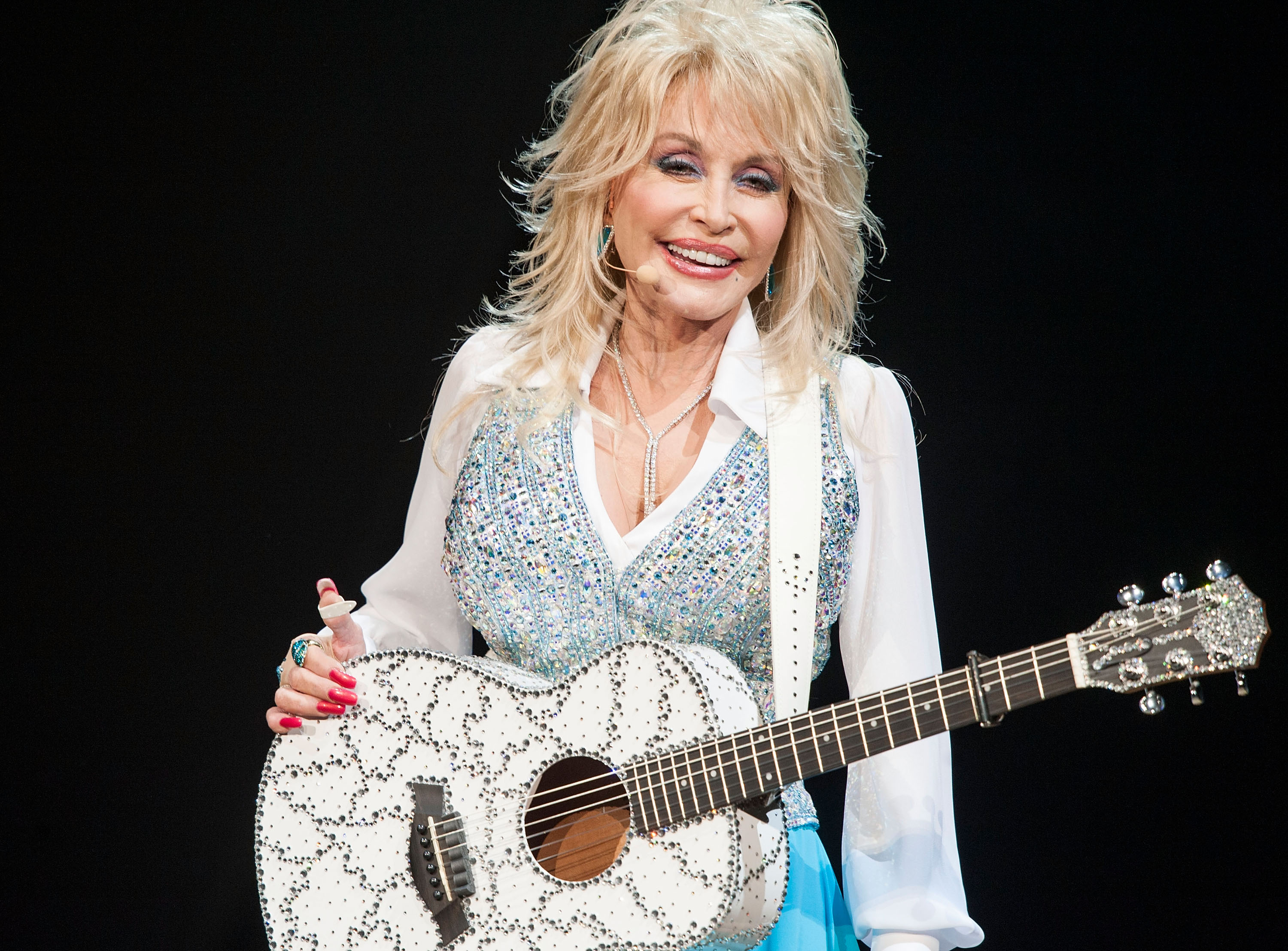 Dolly Parton wears a white rhinestoned shirt and holds a white guitar. 