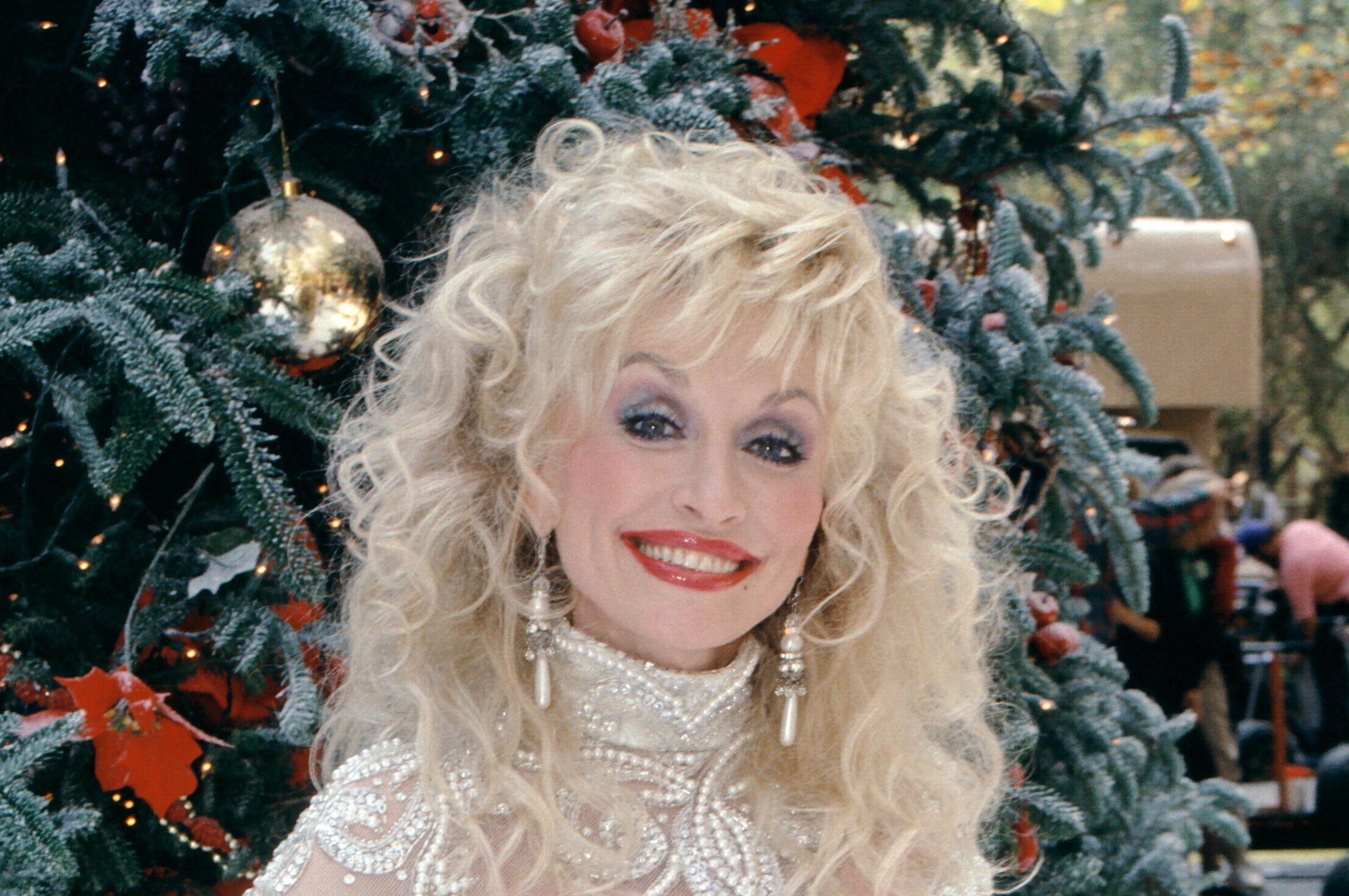 Dolly Parton wears a white beaded shirt and stands in front of a Christmas tree. Dolly Parton's Christmas movies are a part of her holiday themed works.