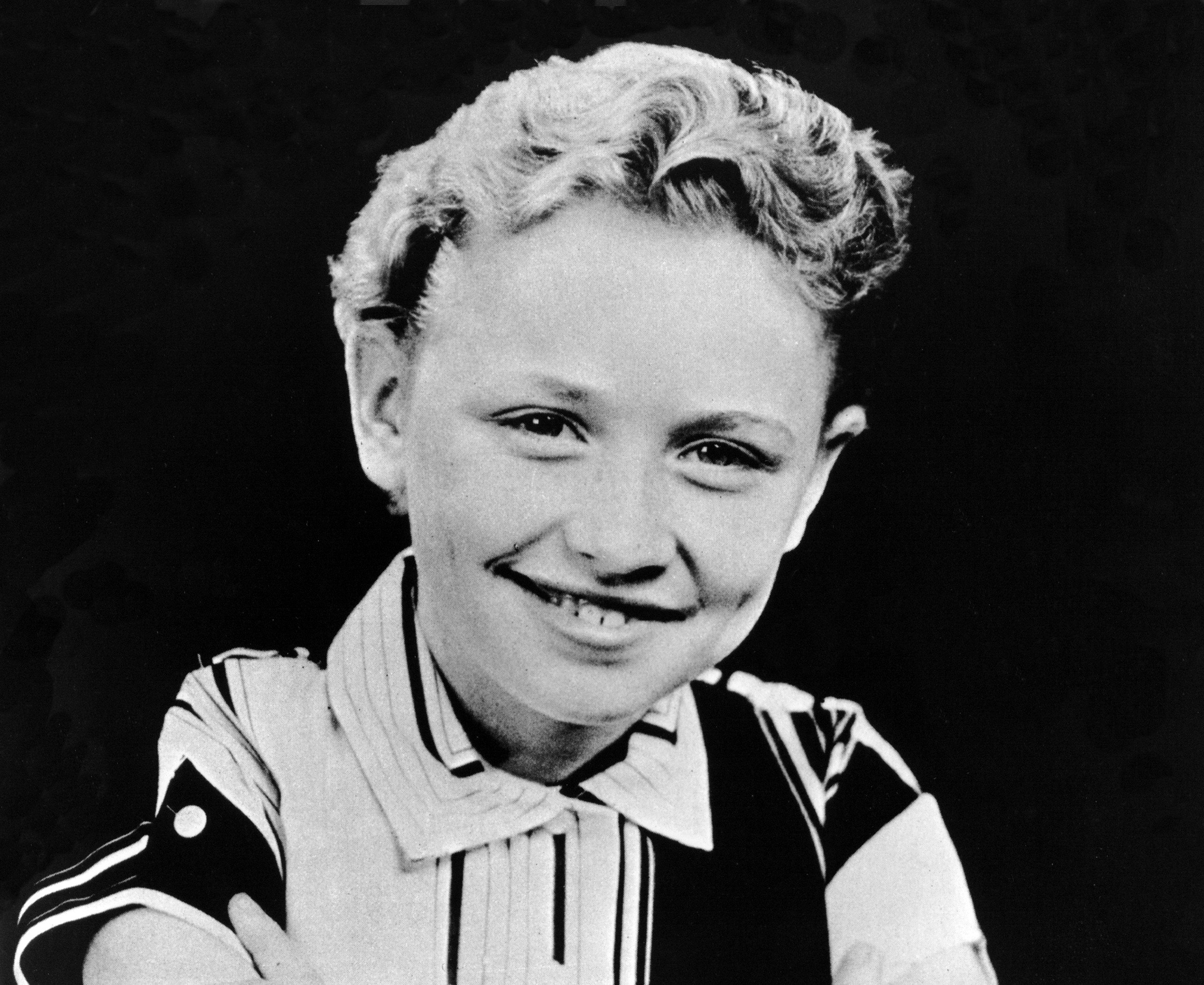 A black and white picture of Dolly Parton as a child. She wears a black and white striped shirt and sits against a black background.