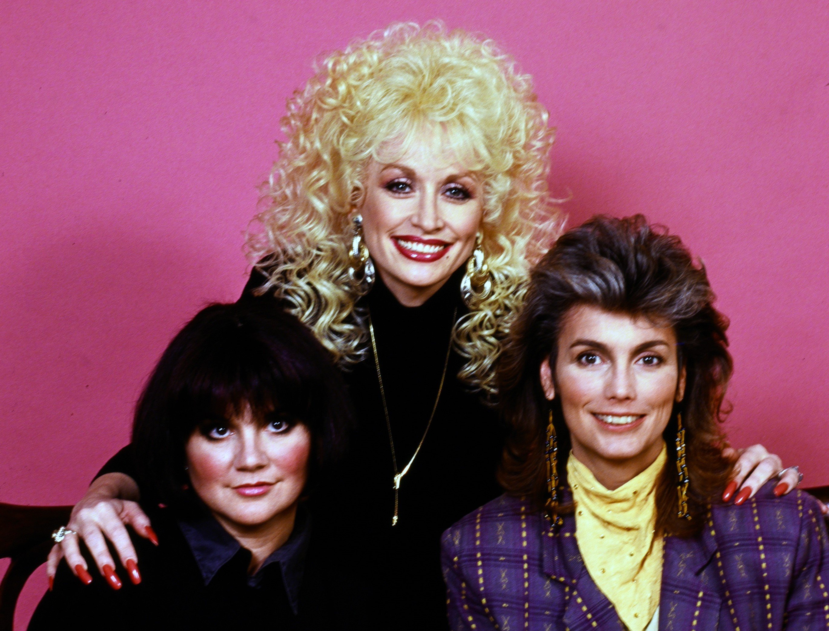 Linda Ronstadt, Dolly Parton, and Emmylou Harris sit against a pink background.