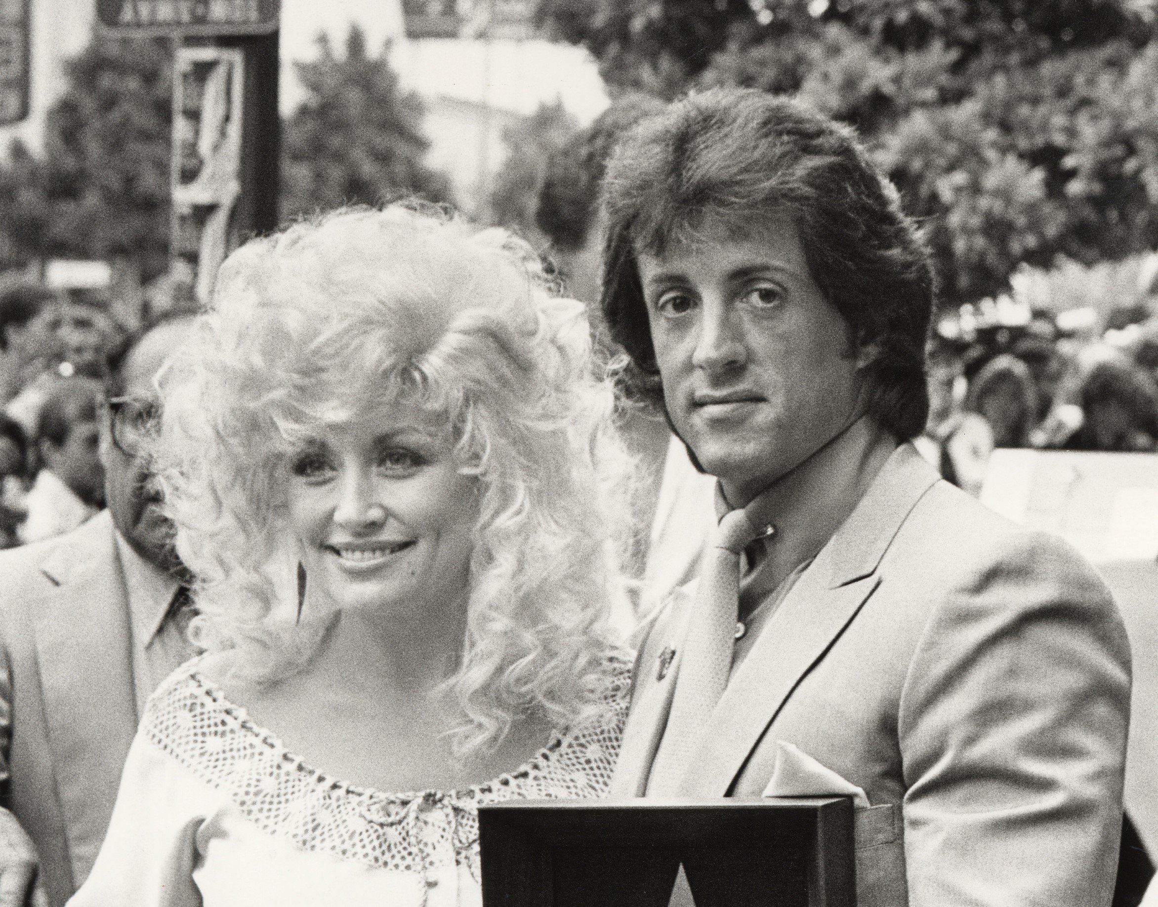 A black and white picture of Dolly Parton wearing a white dress and Sylvester Stallone wearing a suit.