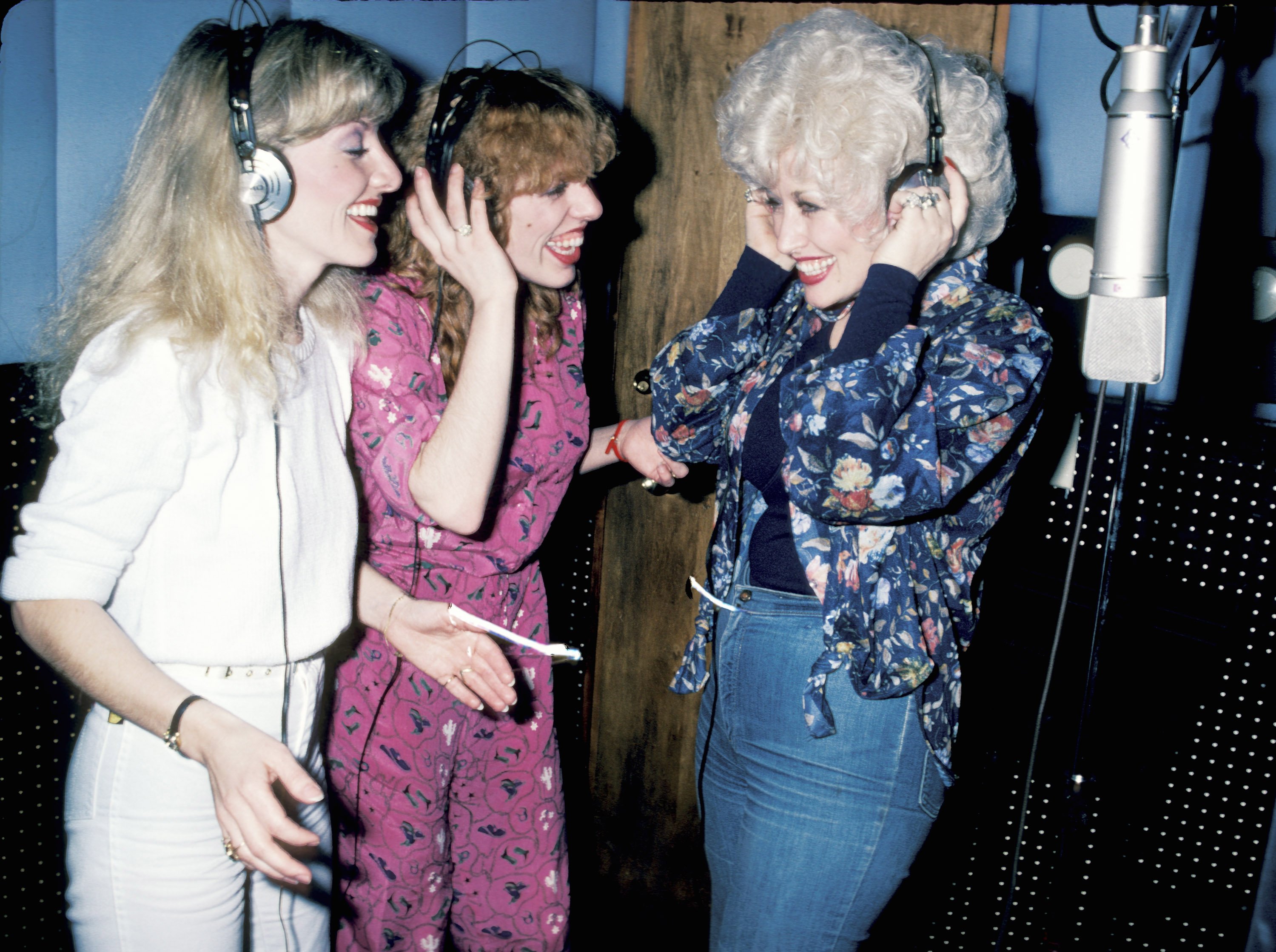 Stella Parton wears a white outfit, Frieda Parton wears a pink outfit, and Dolly Parton wears a blue outfit. They're in the recording studio together.