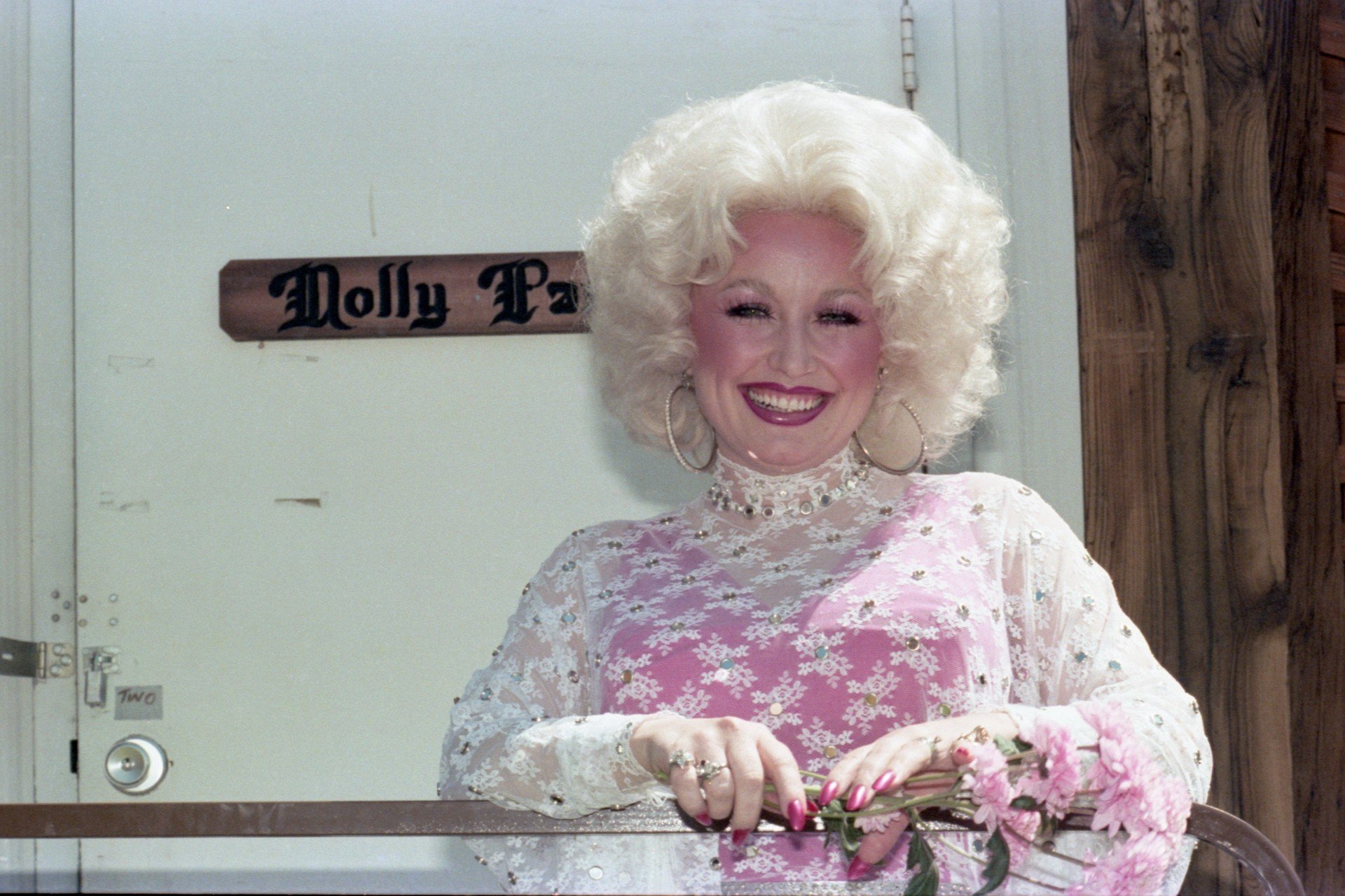 Dolly Parton smiles while wearing a pink and white dress and standing in front of her trailer.