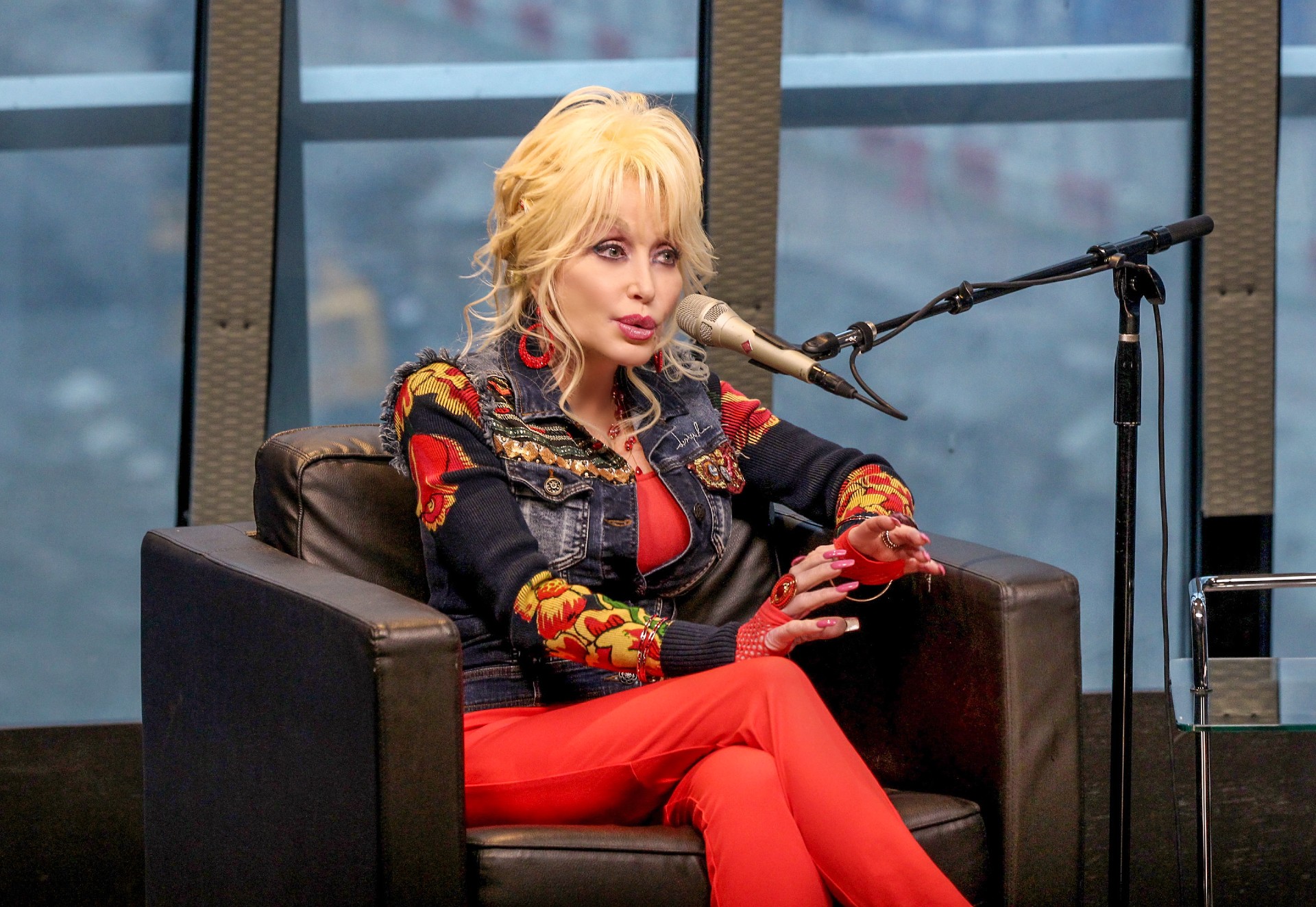 Dolly Parton sits on a couch while talking into a microphone and wearing a red outfit
