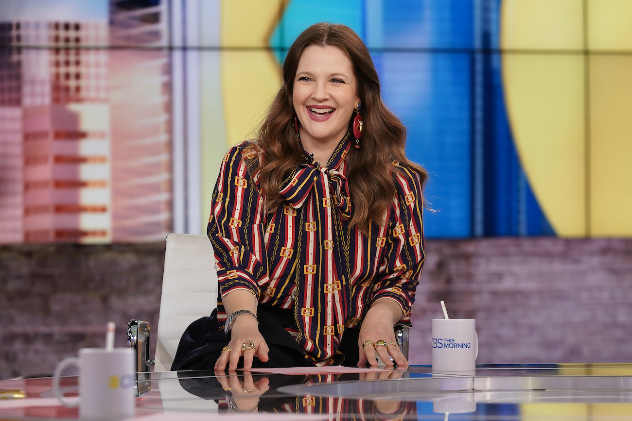 Drew Barrymore smiles as she sits at an anchor desk wearing a striped shirt