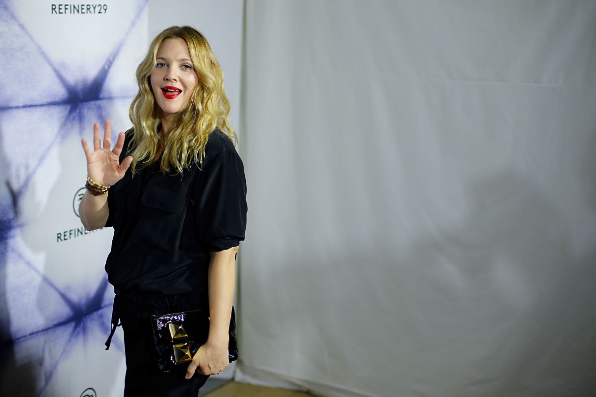 Drew Barrymore in black, holding a clutch in one hand and waving with the other
