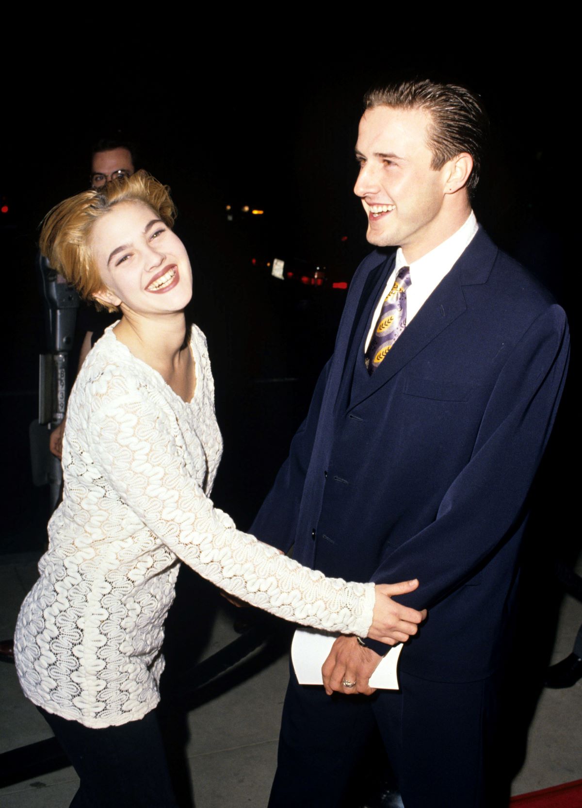 Drew Barrymore smiling a white sweater, reaching out to David Arquette, dressed in a suit