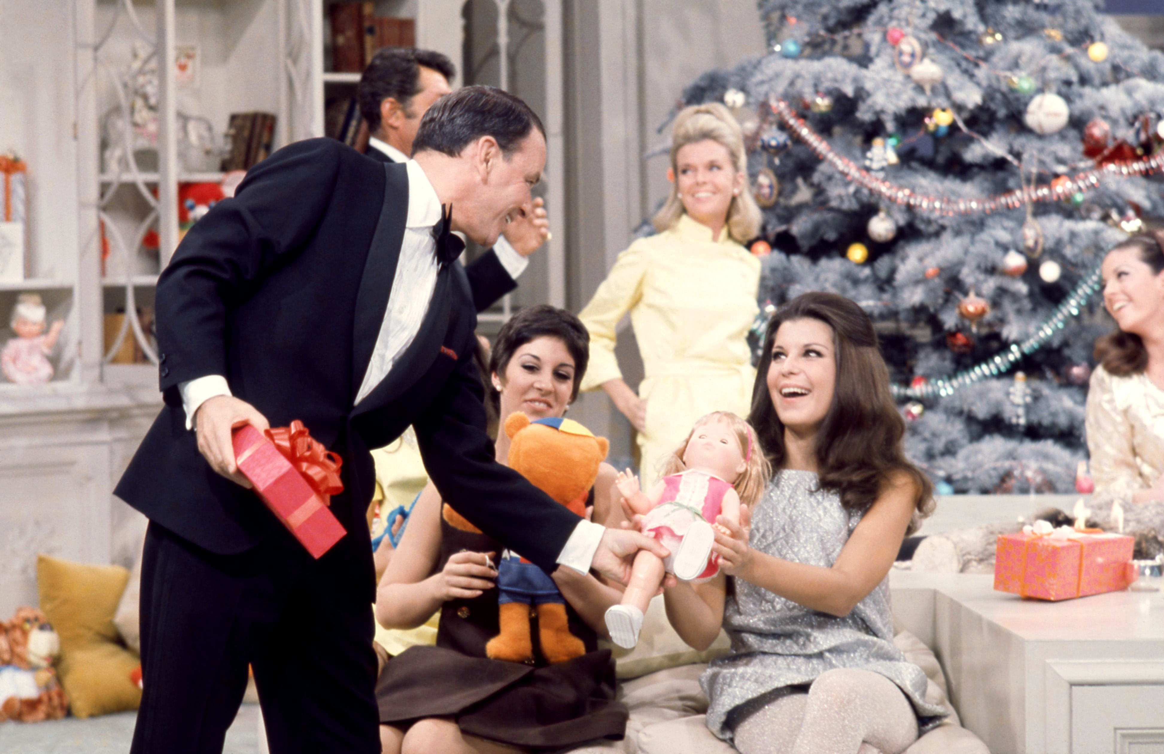 Frank Sinatra wears a tuxedo and hands presents to Deana Martin and Tina Sinatra in front of a Christmas tree.