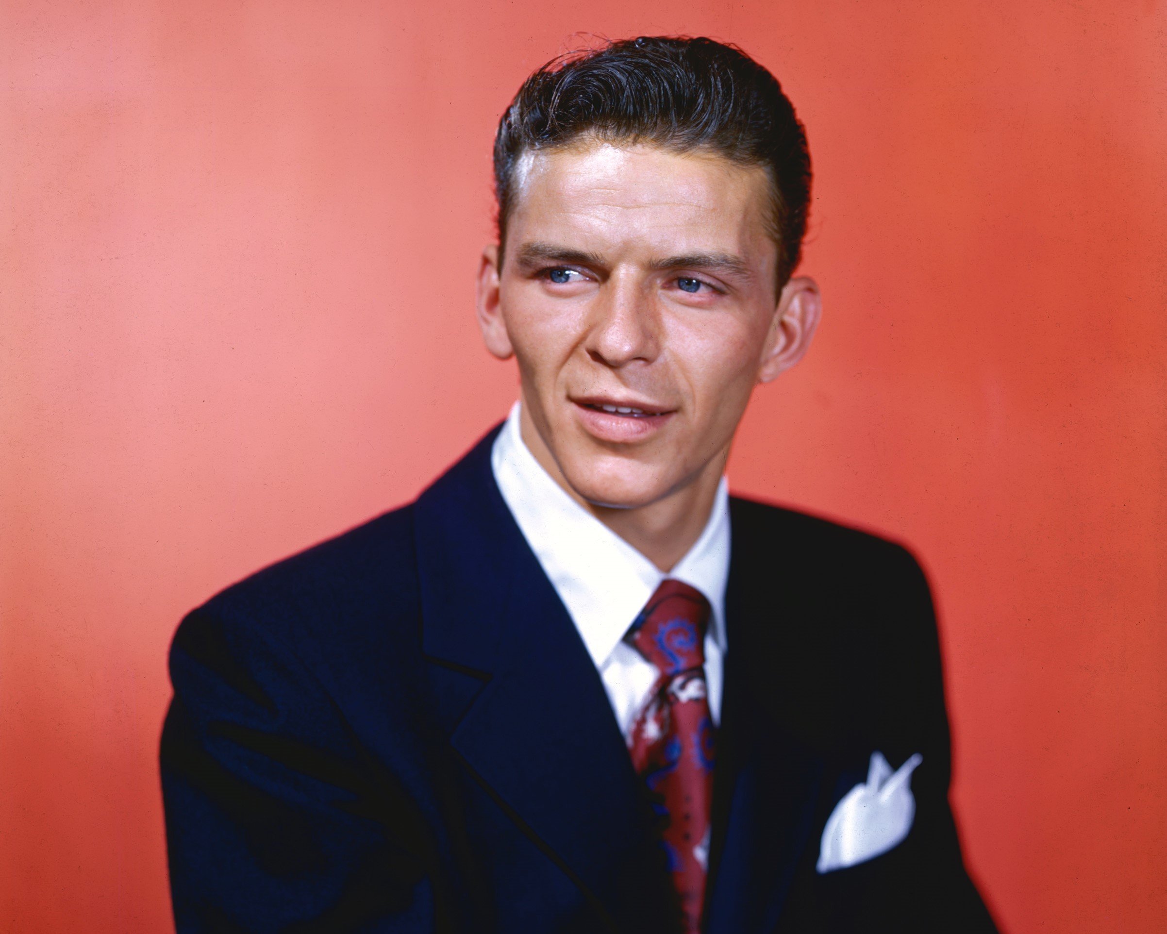 Frank Sinatra wears a suit and sits in front of a red background.