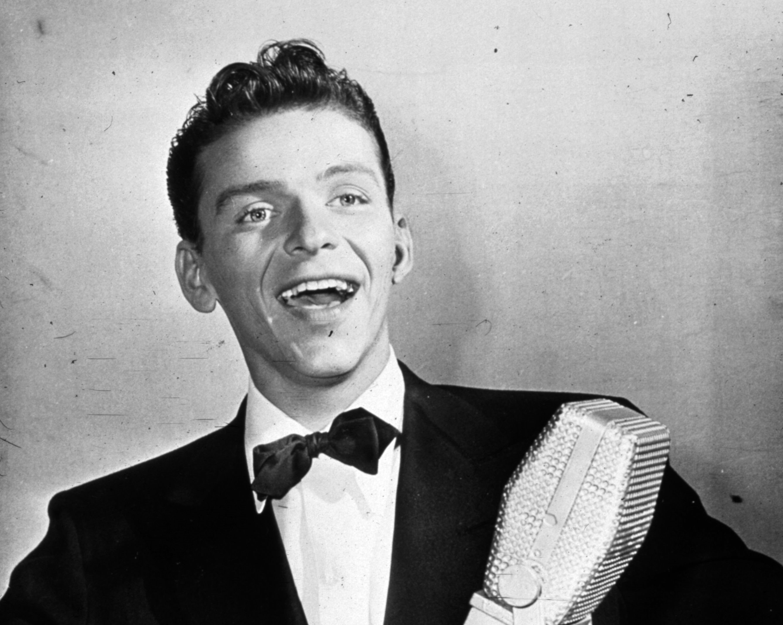 Frank Sinatra wears a tuxedo and stands in front of a microphone.
