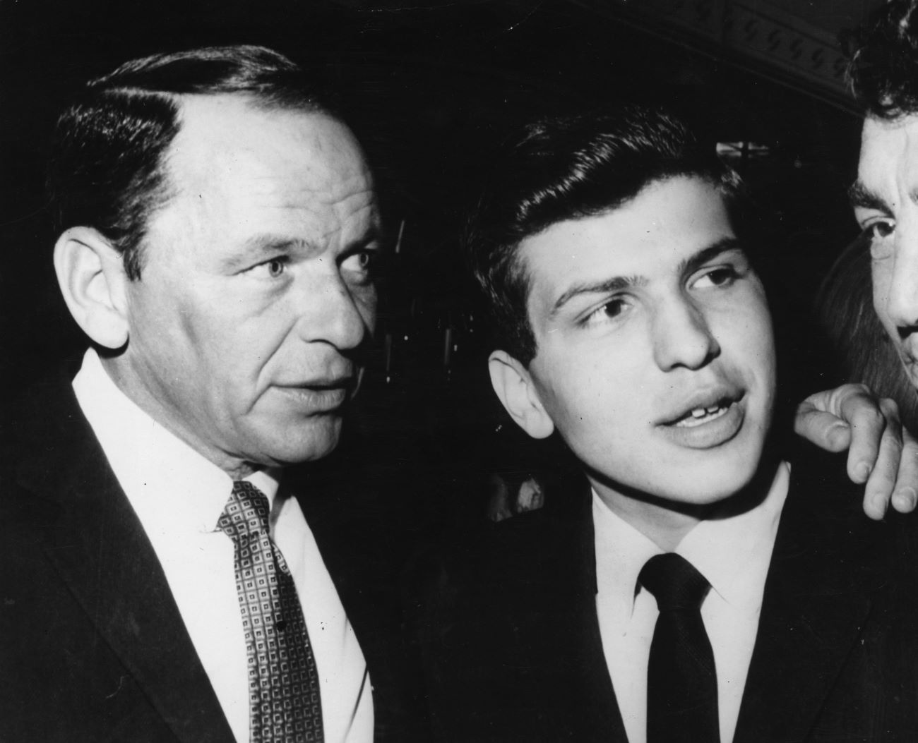 Frank Sinatra stands with his arm around the shoulders of Frank Sinatra Jr. They both wear suits.