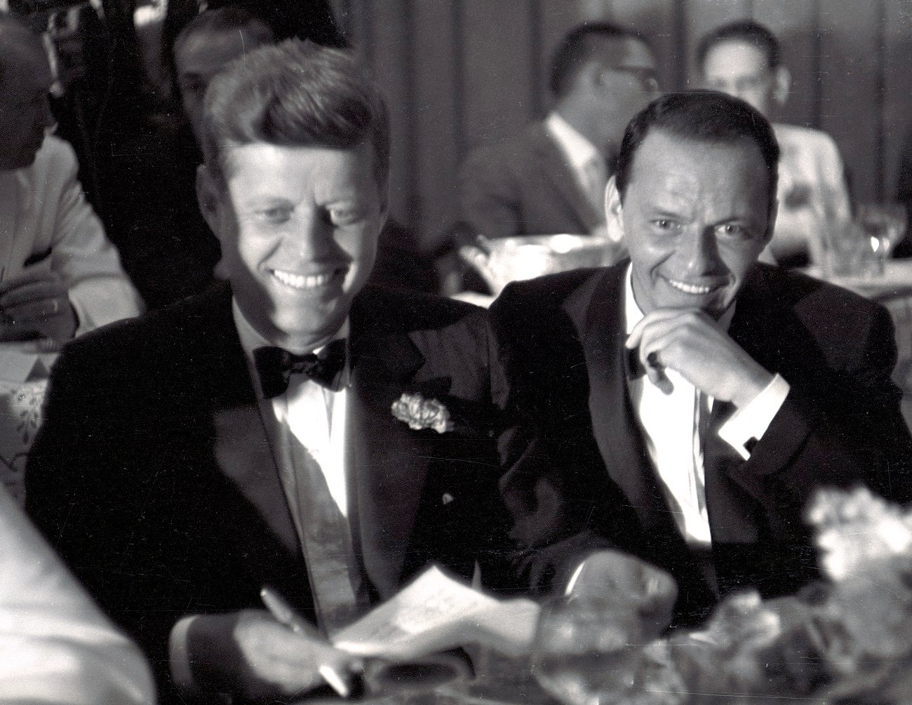 A black and white photo of John F. Kennedy and Frank Sinatra wearing tuxedos and sitting next to each other.