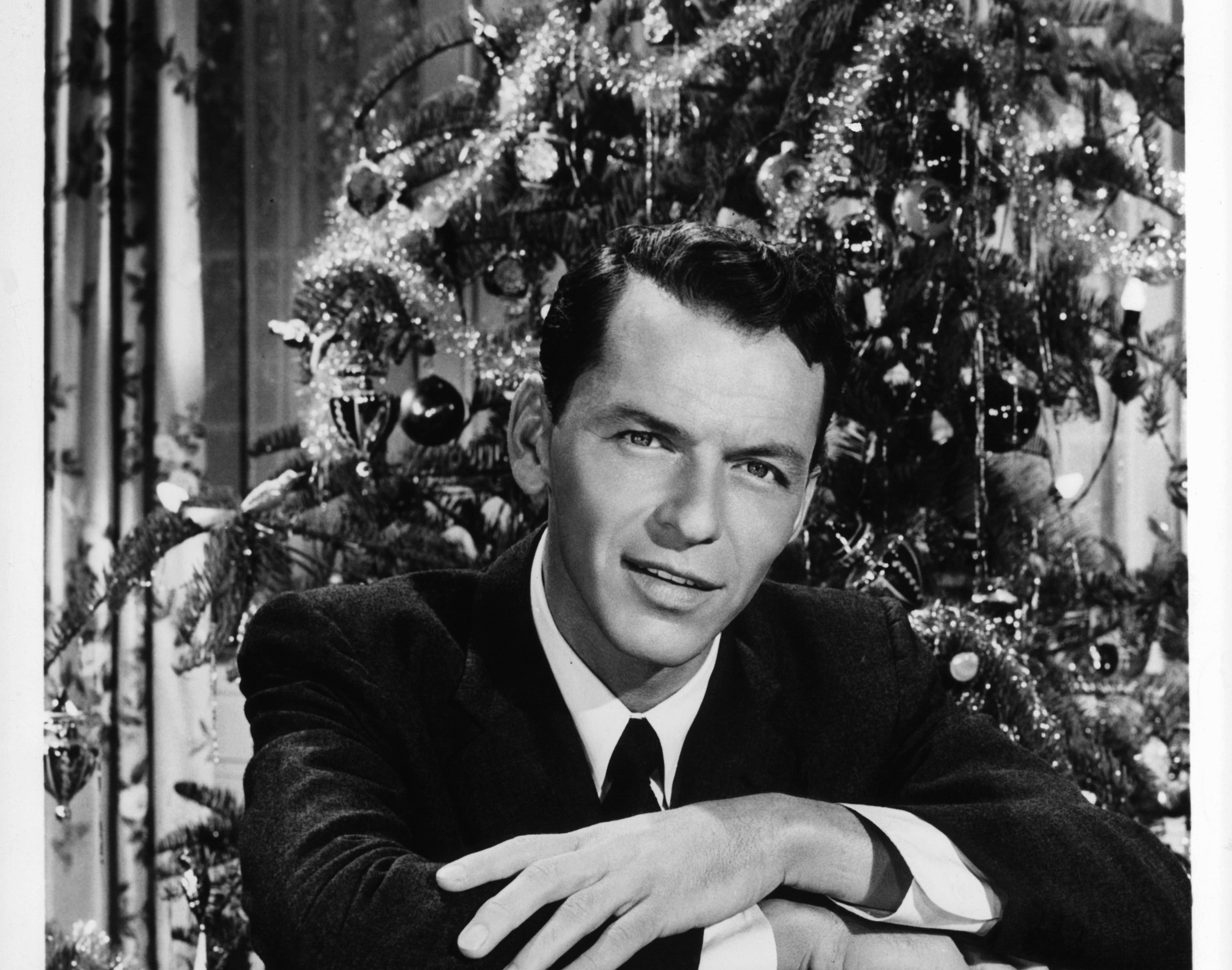 Frank Sinatra wears a suit and sits in front of a Christmas tree.